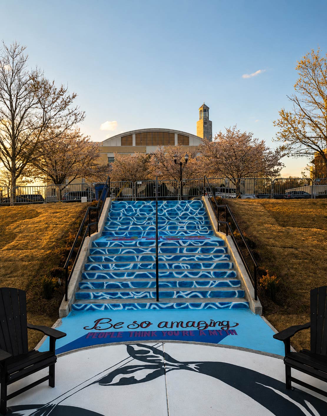 Staircase with blue design and text, flanked by benches, leading to a building with trees and a clear sky in background.