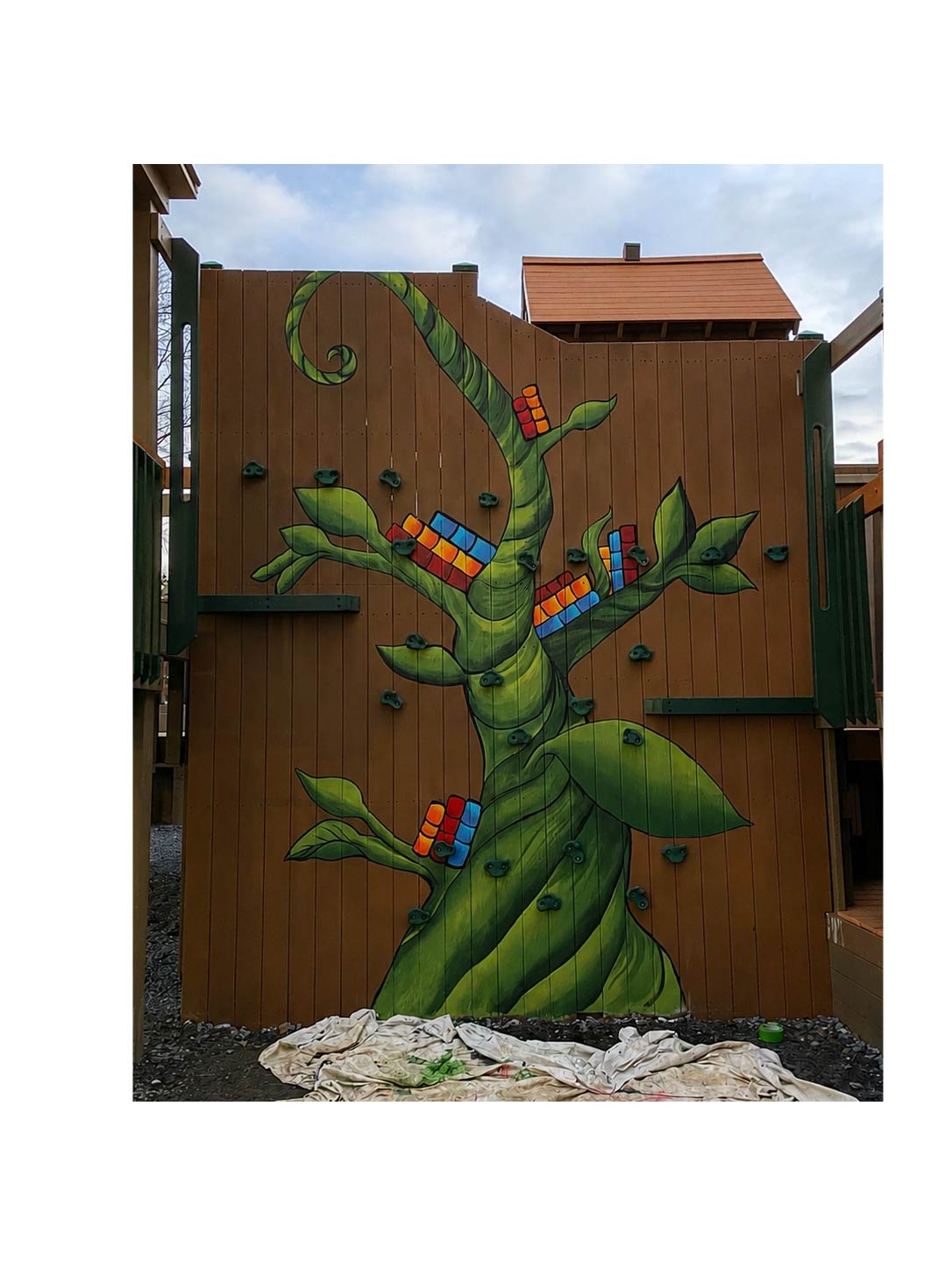 Colorful painted tree with toys on branches, set against a wooden fence and sky background.