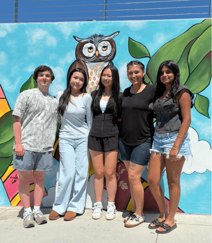 Five people stand in front of a colorful mural with an owl and leaves, smiling outdoors on a sunny day.