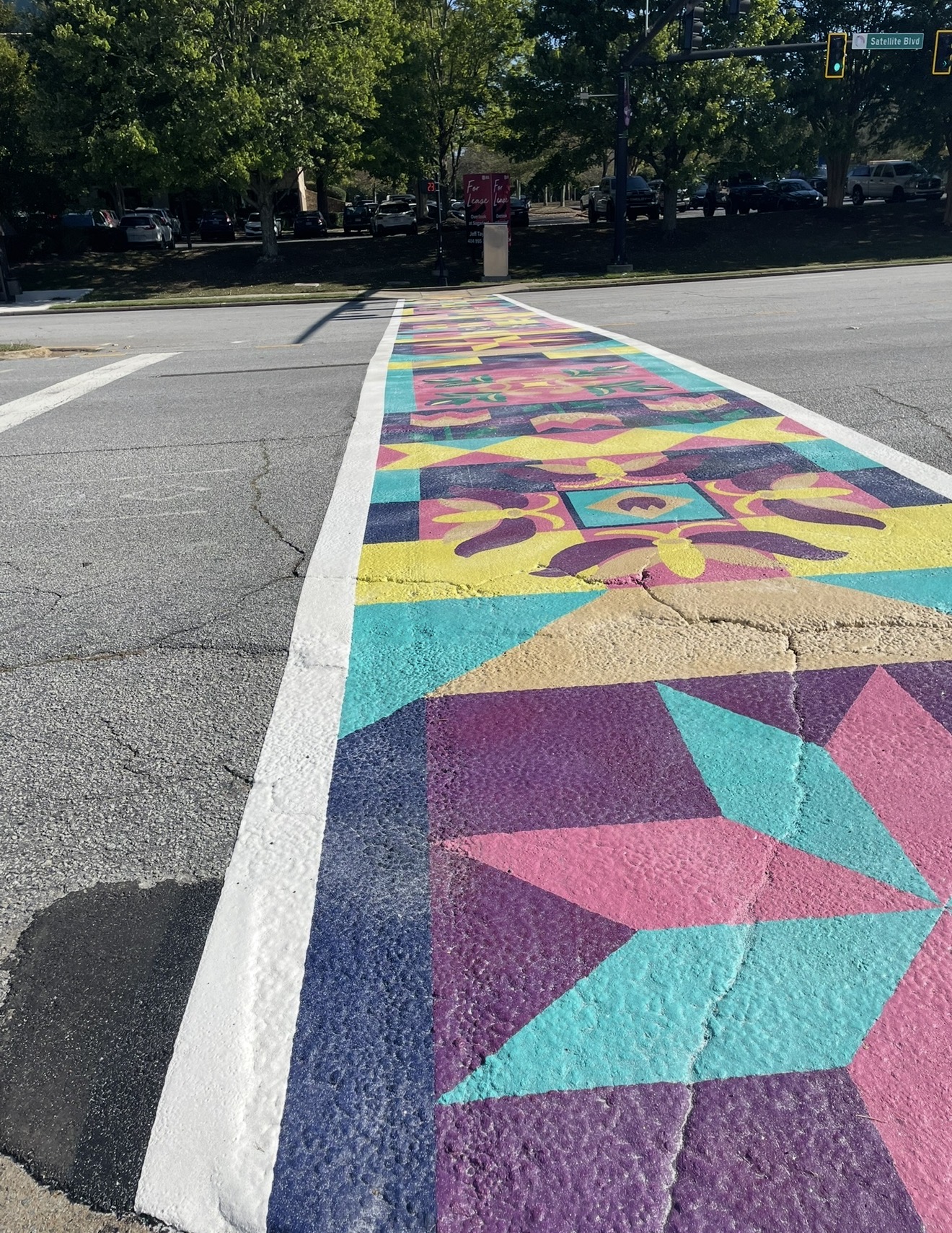 Colorful geometric mural on street crosswalk with trees and cars in background.