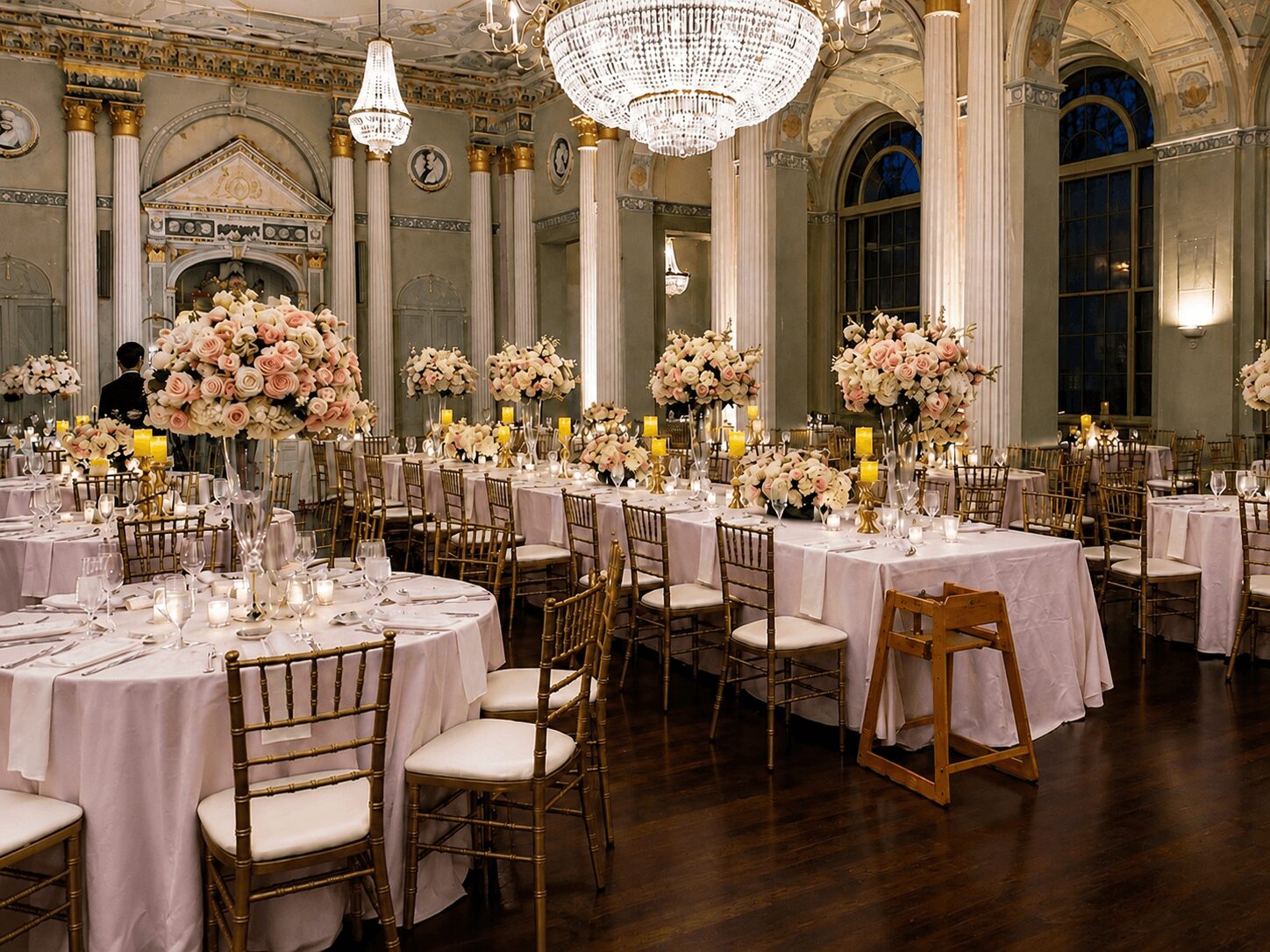 Elegant banquet hall with round tables, floral centerpieces, chandeliers, and tall windows, set for a formal event.