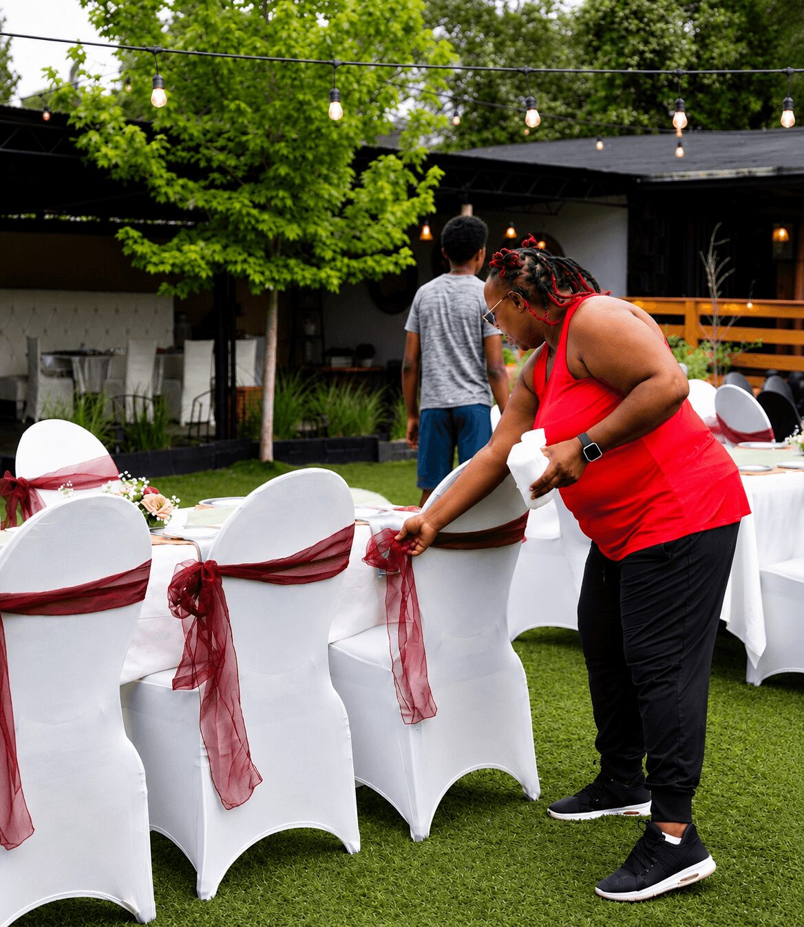 Woman in red top decorating a white chair with a red sash at an outdoor event, with tables and string lights in background.