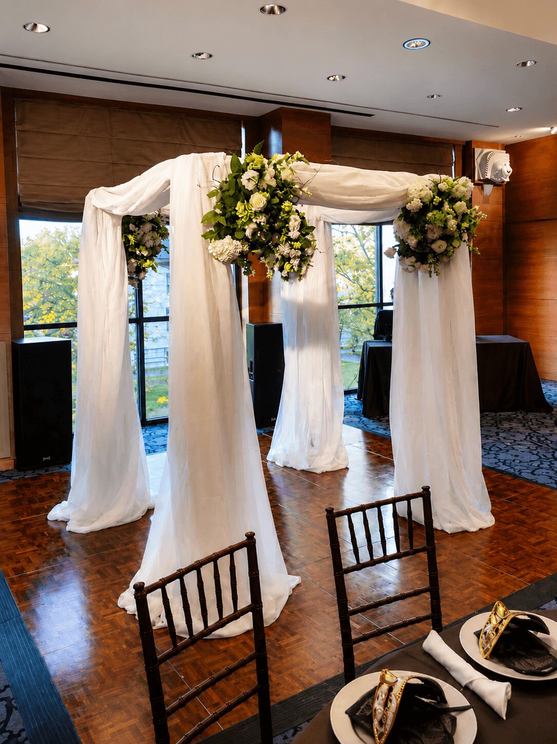 Wedding arch decorated with white fabric and floral arrangements, set indoors with windows and tables nearby.