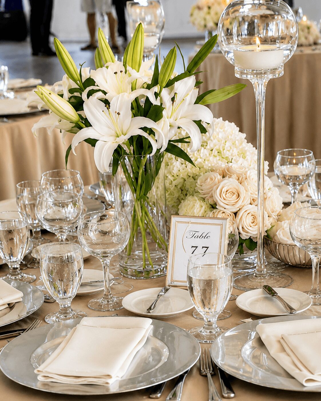 Elegant table setting with white lilies, roses, glassware, and a table number card, in a formal event setting.