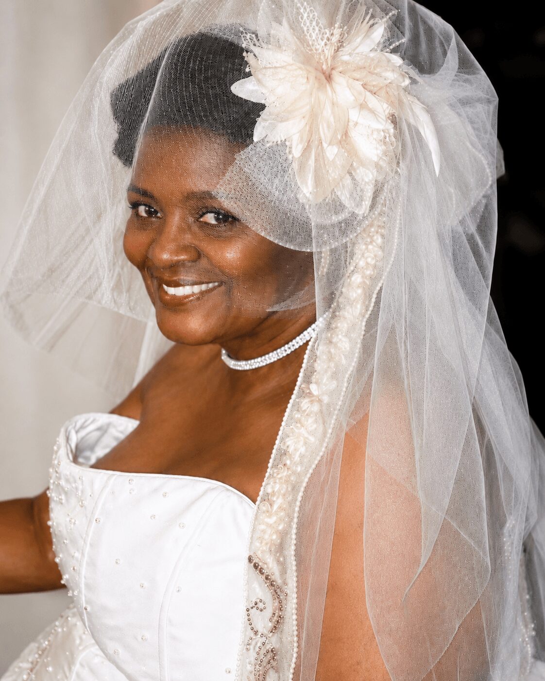 Smiling woman in wedding dress with veil, large flower headpiece, and pearl necklace, looking at camera.