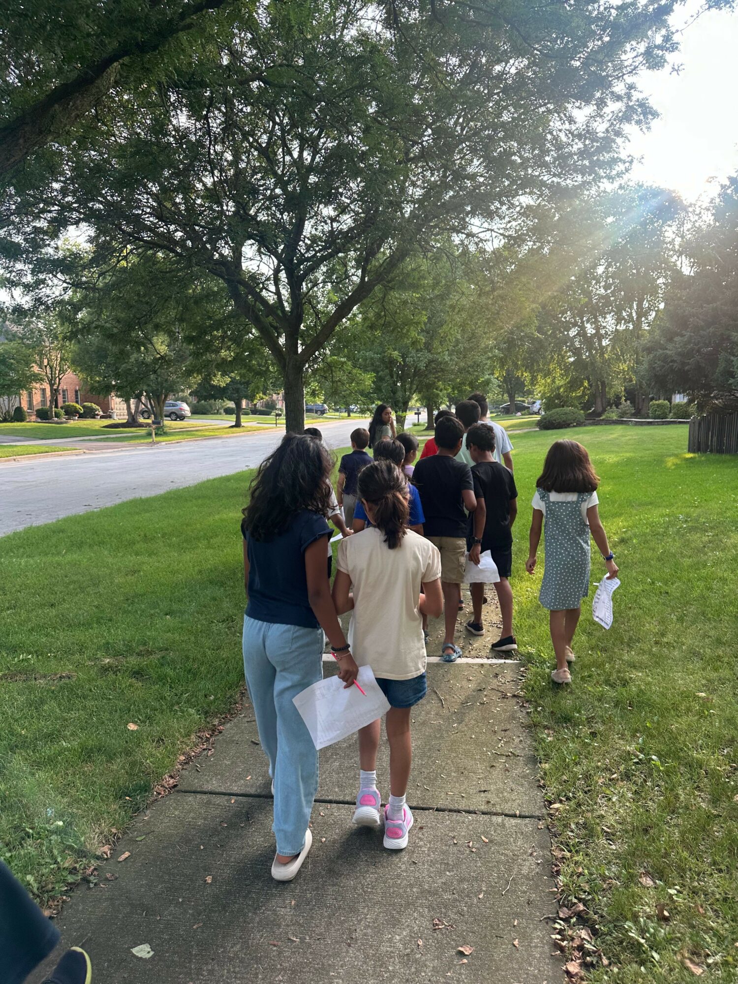 Group of people walking on a sidewalk in a park with trees and grass, some carrying bags or papers.