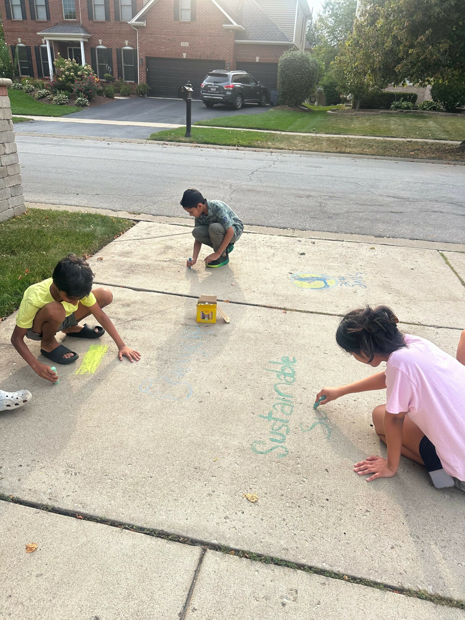 Two children playing on a sidewalk with chalk, one crouching and the other sitting, near a grassy area and street.