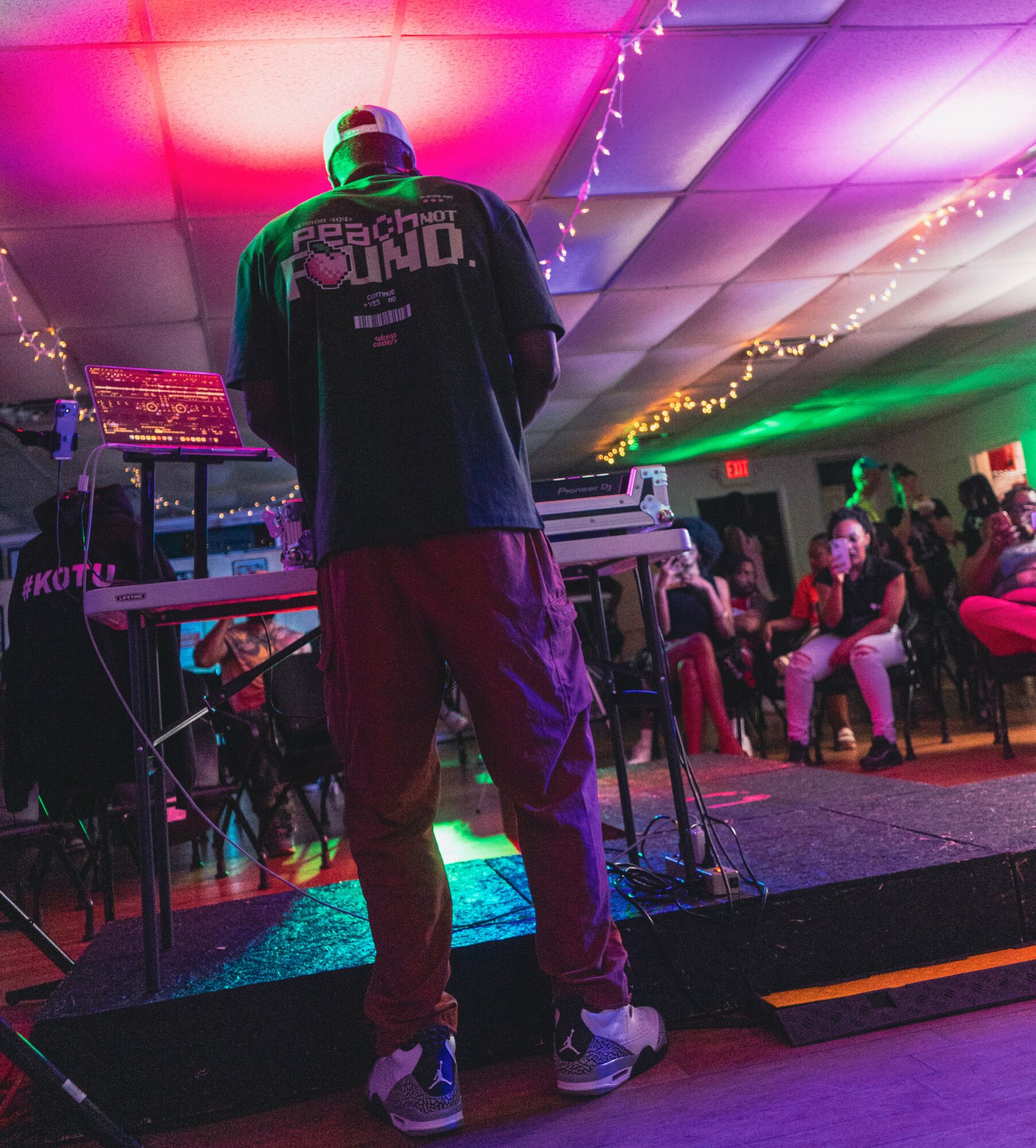 Person standing on stage with DJ equipment, colorful ceiling lights, and audience seated in background.