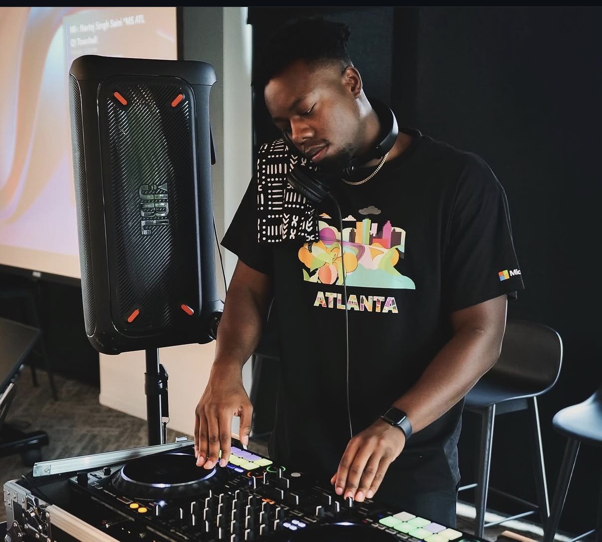 DJ with headphones operating a mixer, standing next to a speaker in a dark room, wearing a black T-shirt with colorful graphics and 'ATLANTA' text.