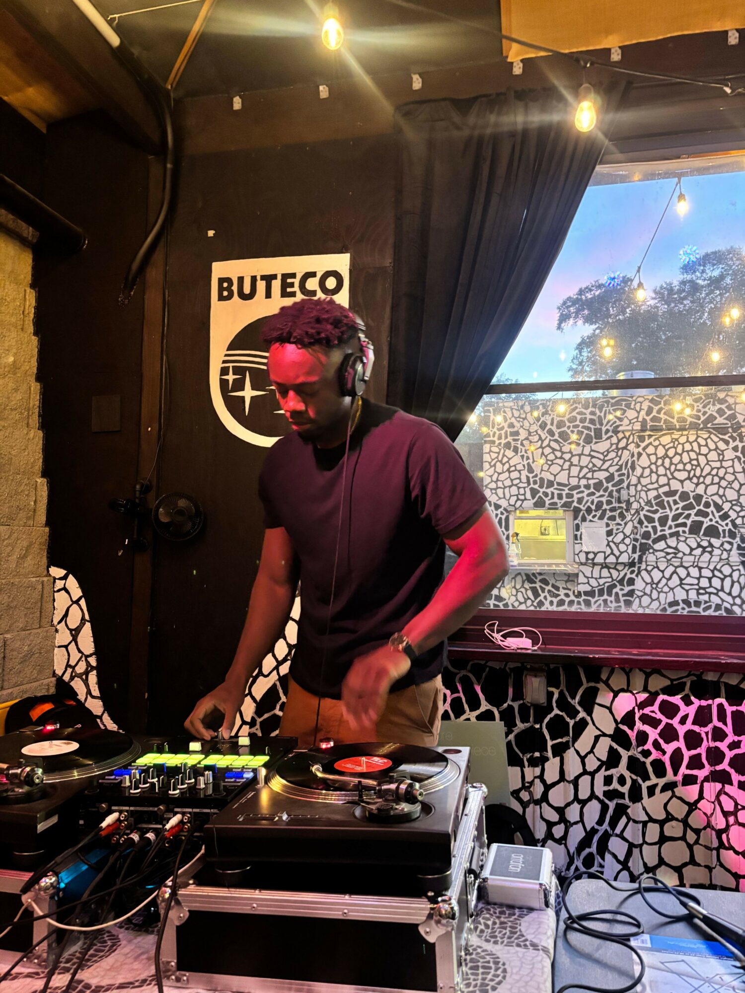 Person DJing with headphones, standing at turntables in a decorated indoor space, with a Buteco sign in the background.