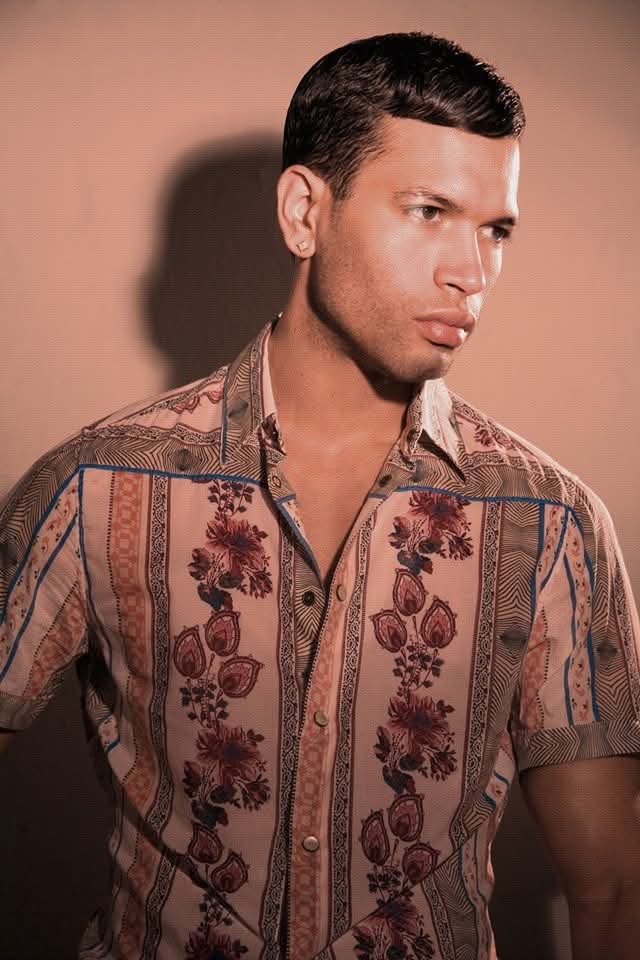 Young man with short dark hair wearing a patterned short-sleeve shirt, looking to the side against a plain background.