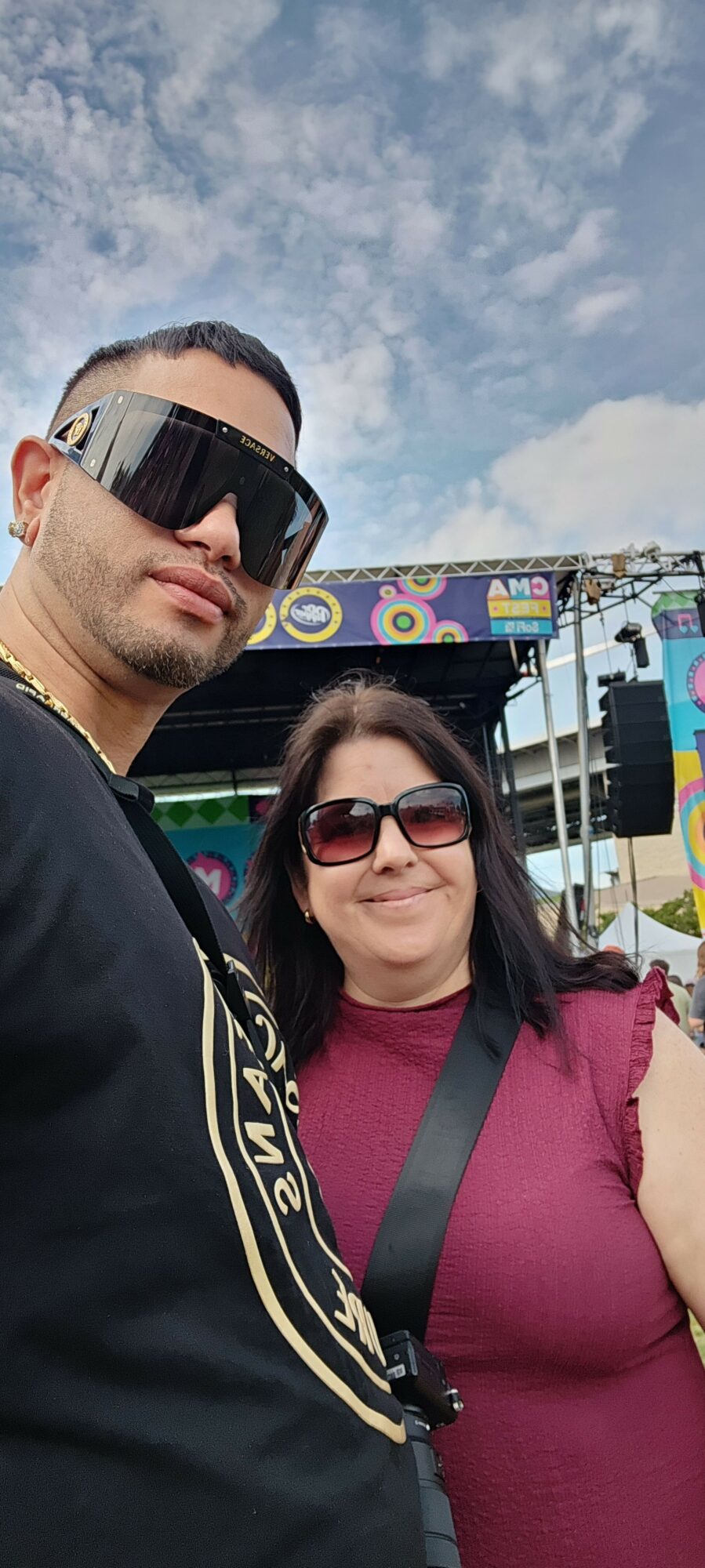 Two people wearing sunglasses, one with a camera, outdoors with a cloudy sky background.