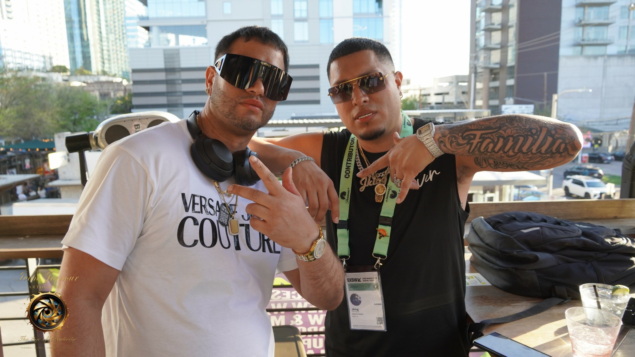 Two men pose indoors with city buildings visible outside. One wears sunglasses and a white shirt, the other wears glasses and a black shirt with tattoos.