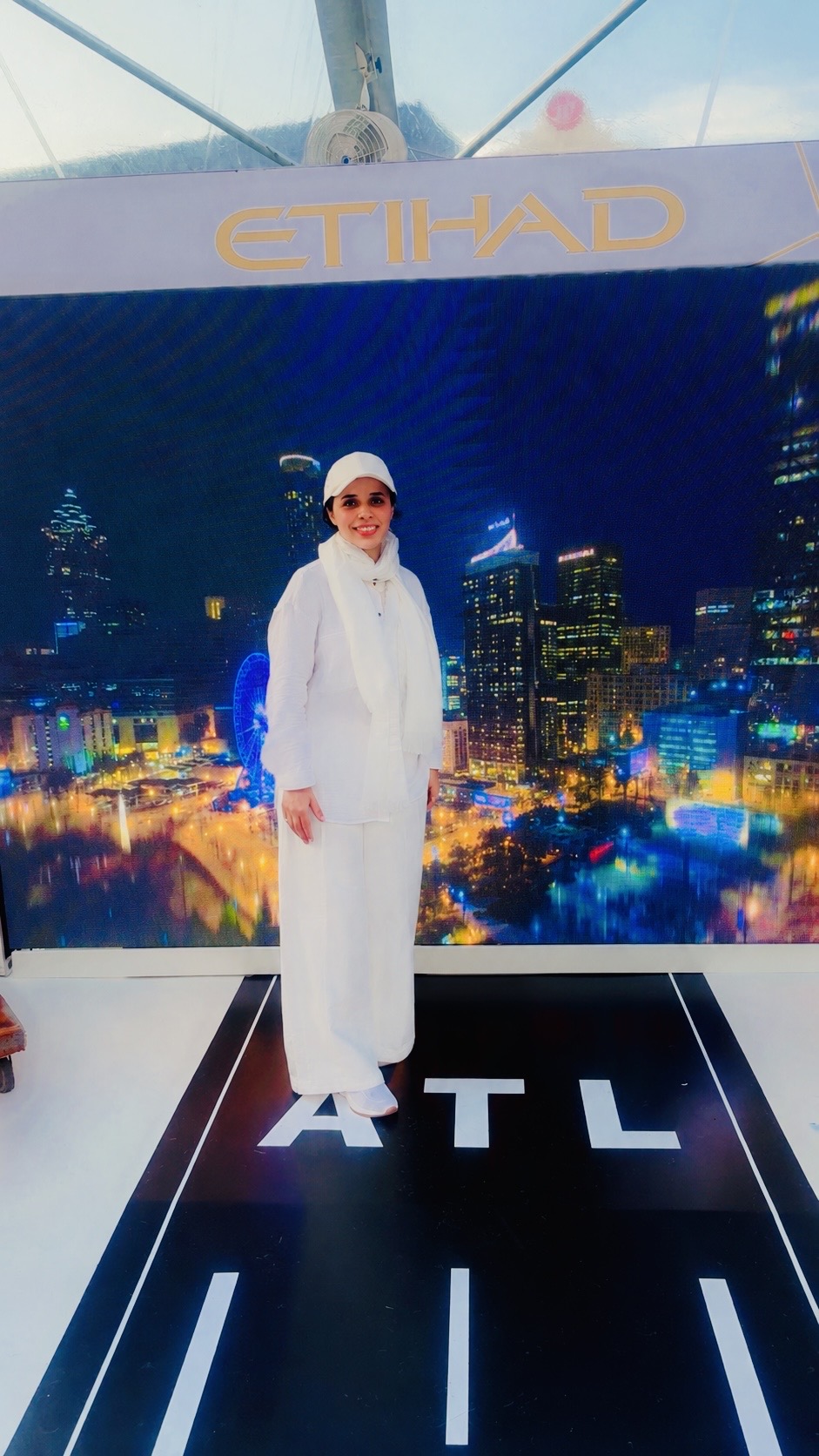 Person in white outfit and sunglasses standing in front of a cityscape backdrop with 'ETIHAD' and 'ATL' text.