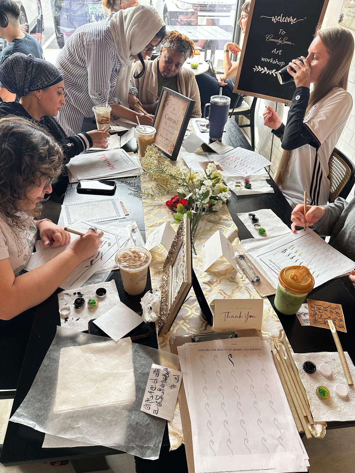 Group of women sitting around a table with drinks, flowers, and notebooks, engaged in conversation and writing, in a cafe setting.