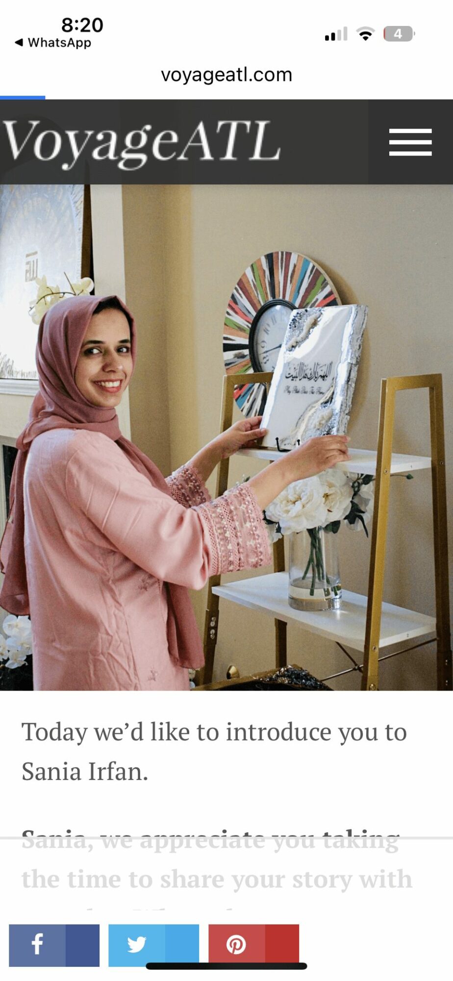 Woman in pink hijab painting on canvas in a bright room with a mirror and flowers, smiling at camera.