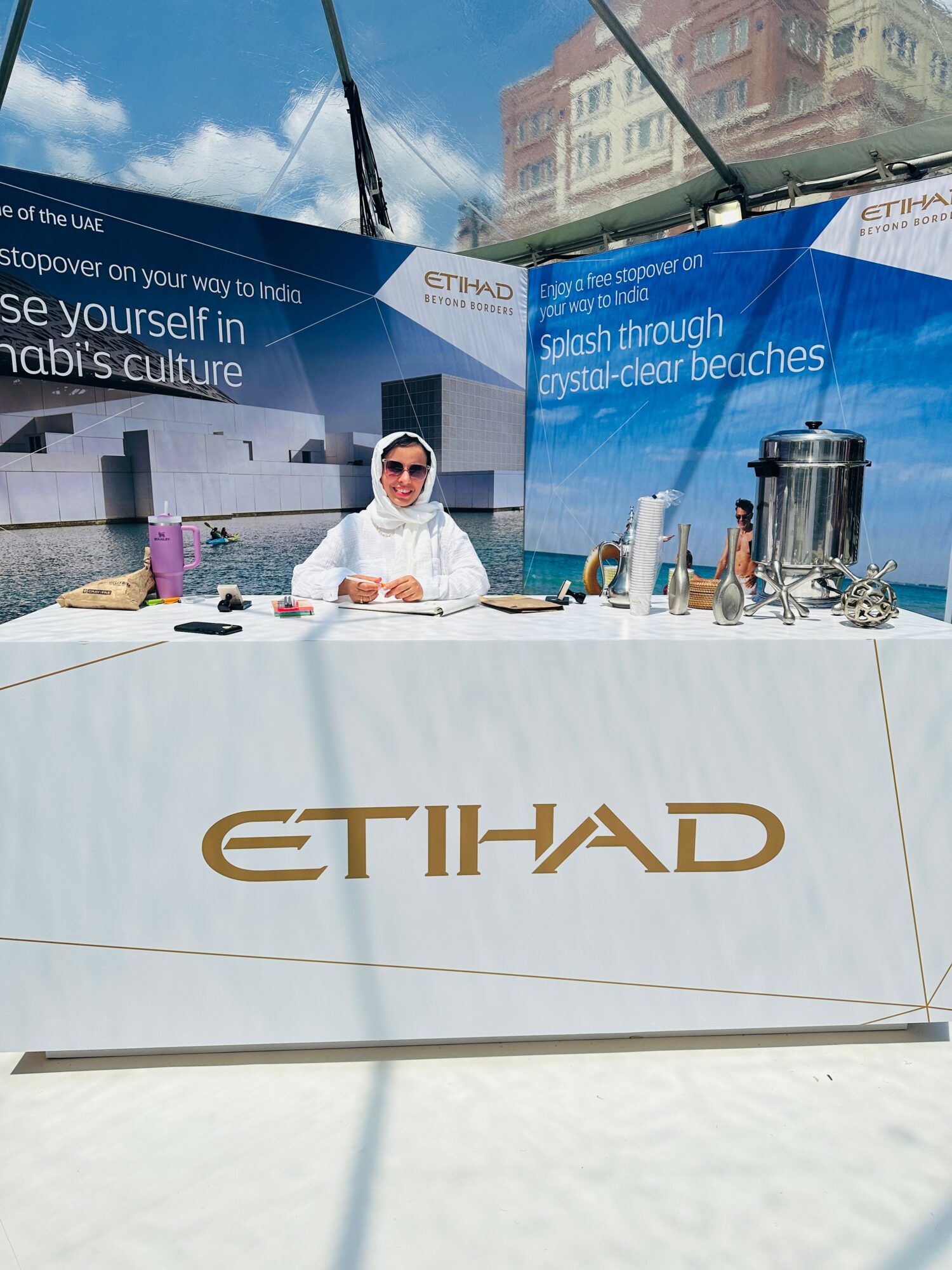 Person in white clothing and sunglasses sitting at a table with ETIHAD branding, outdoor setting with blue sky and buildings