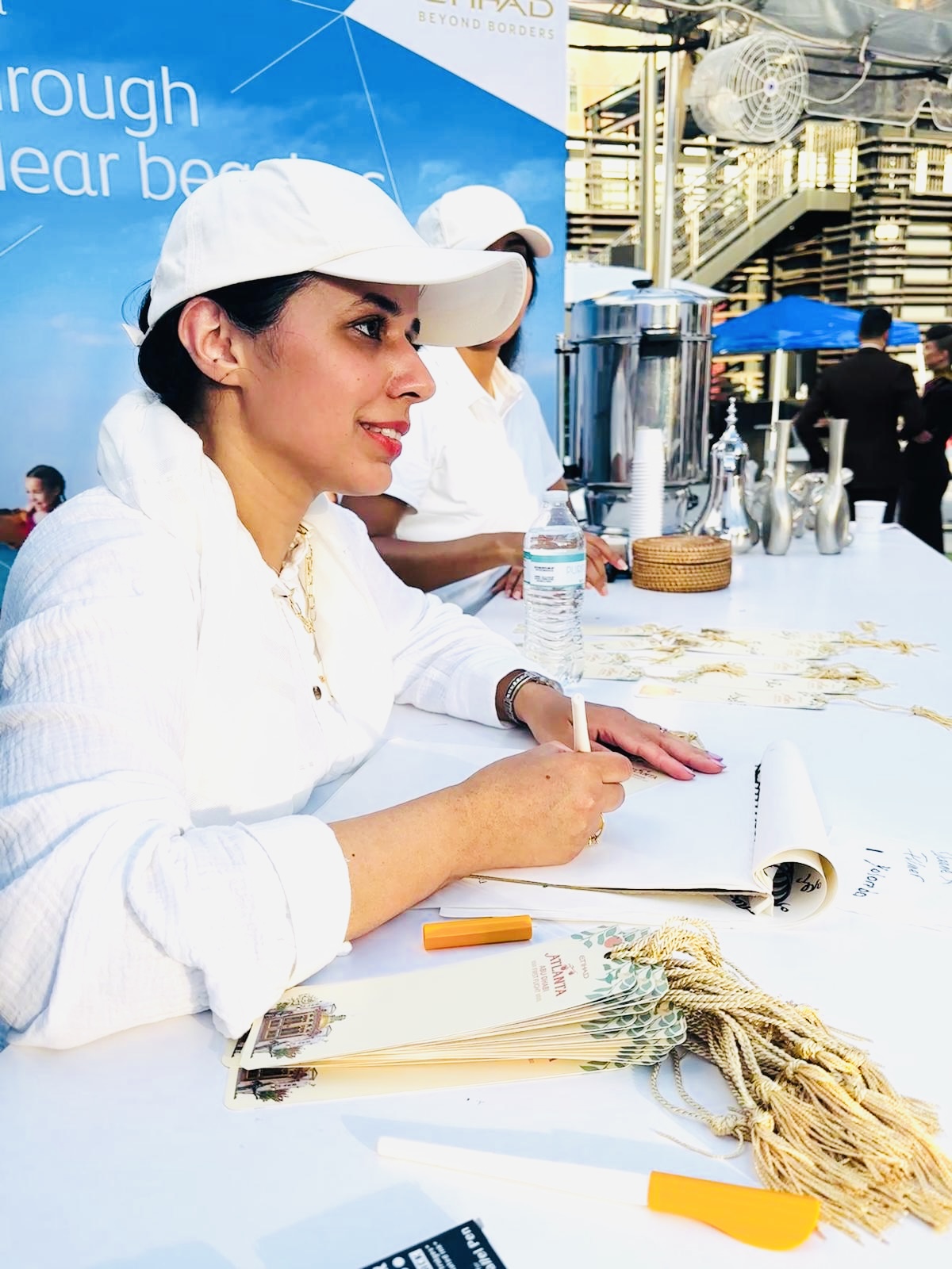 Woman in white cap and shirt sitting at a table outdoors, writing in a notebook with a pen, with a blue background and people in the background.