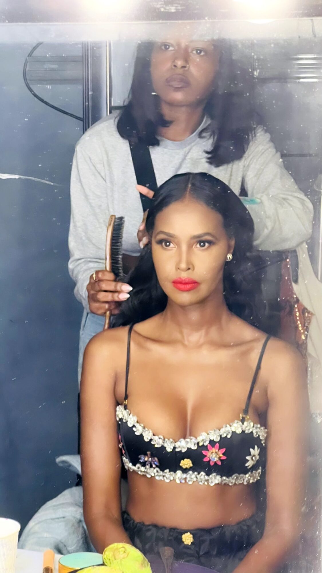 A woman with long dark hair and red lipstick sits in front of a mirror, getting her hair styled by a hairstylist.