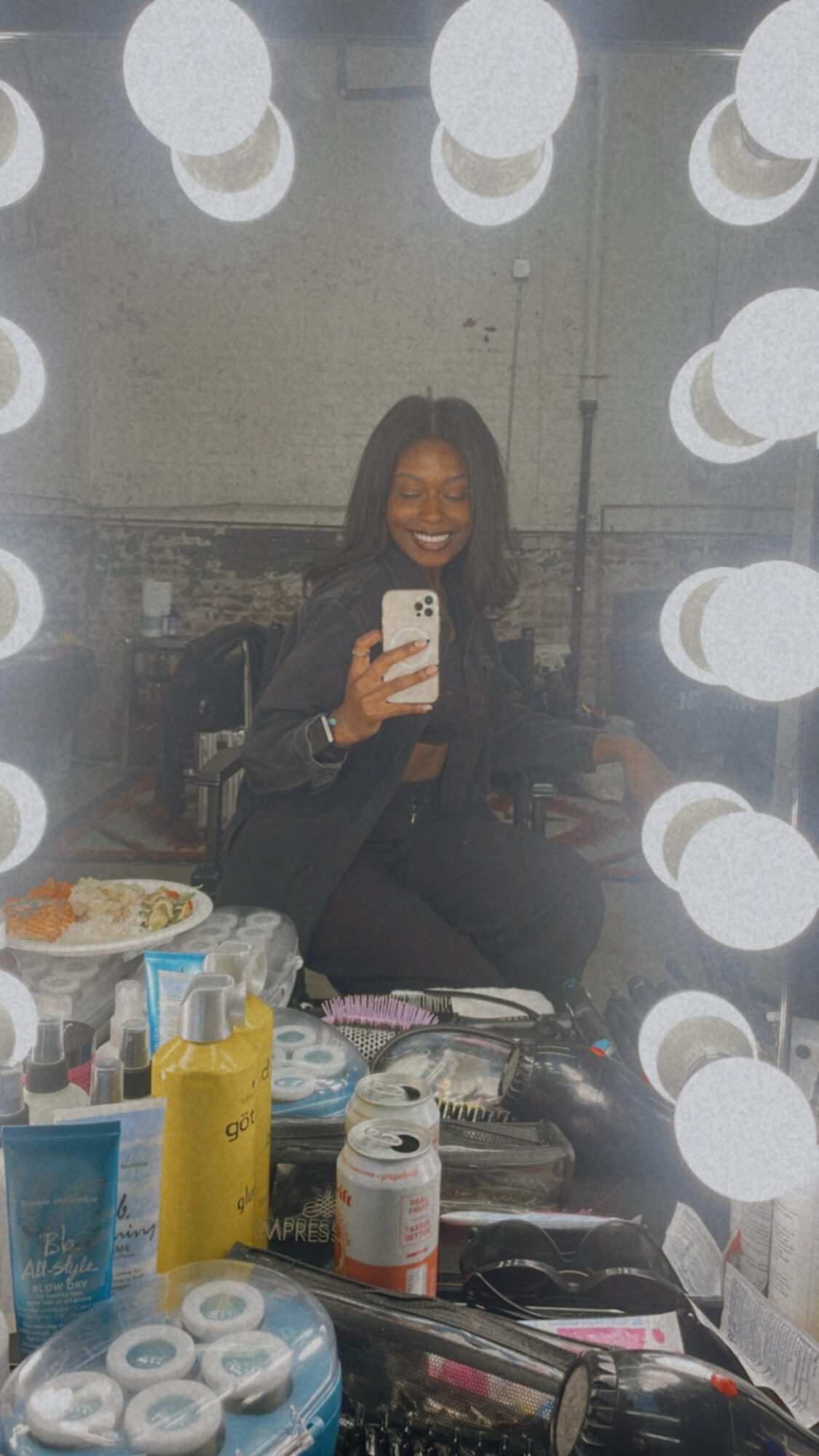 Person taking a selfie in front of a mirror with round lights, smiling, with various items on the table in front.