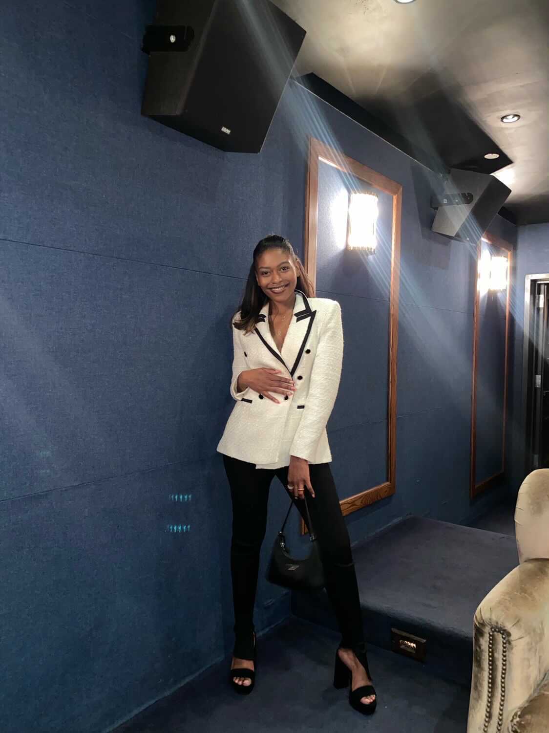 Woman in white blazer and black pants standing in a room with blue walls and speakers, smiling at the camera.