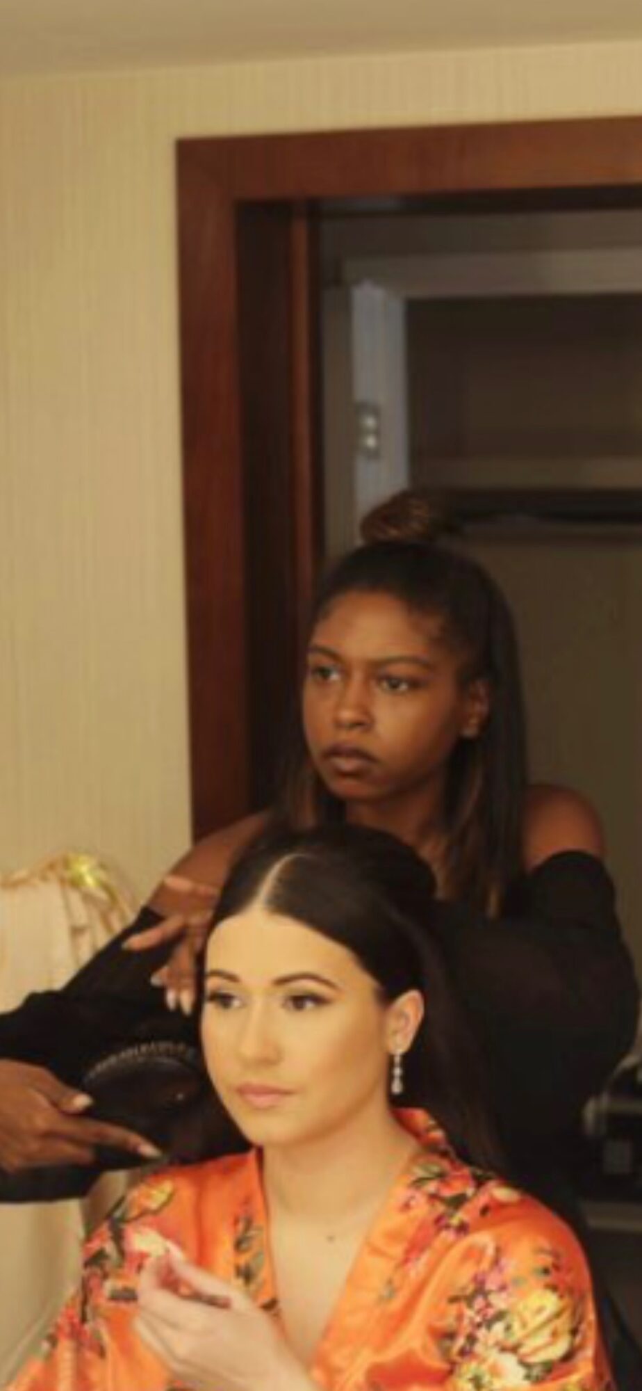 Two women, one with dark hair and one with lighter hair, in a room with a doorway and a mirror behind them.
