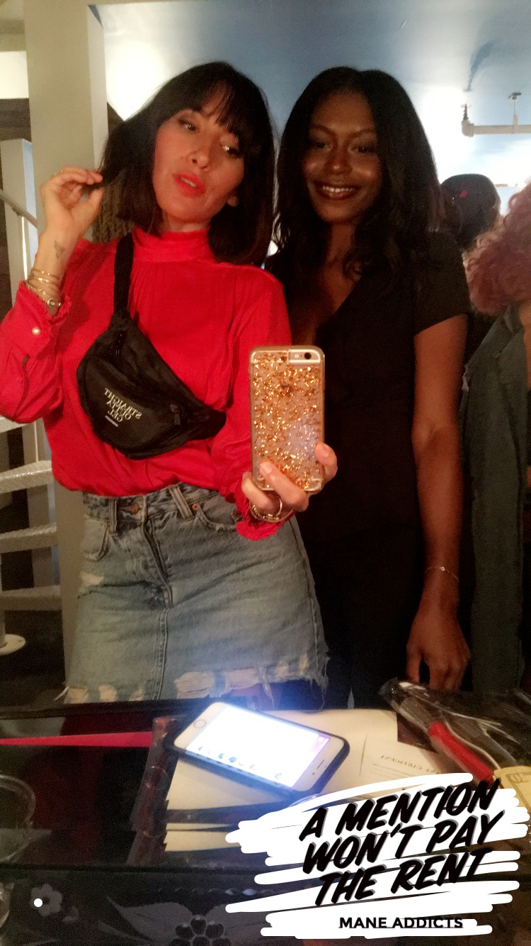 Two women posing for a photo indoors, one in a red top and denim skirt, the other in black, with a phone and a sign on the table.