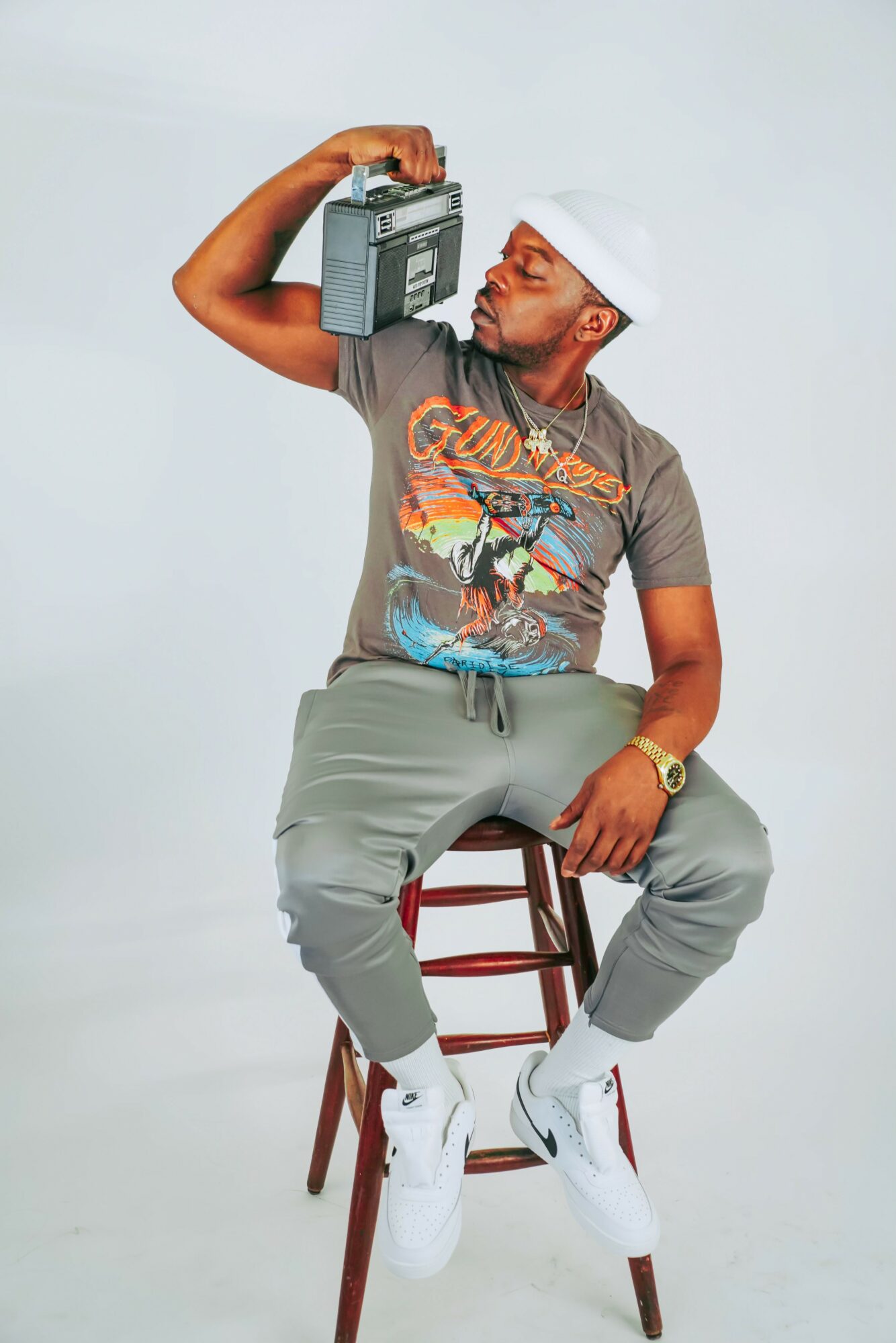 Man sitting on a stool, wearing a white cap, gray t-shirt, gray pants, and white sneakers, holding a boombox, looking at it.