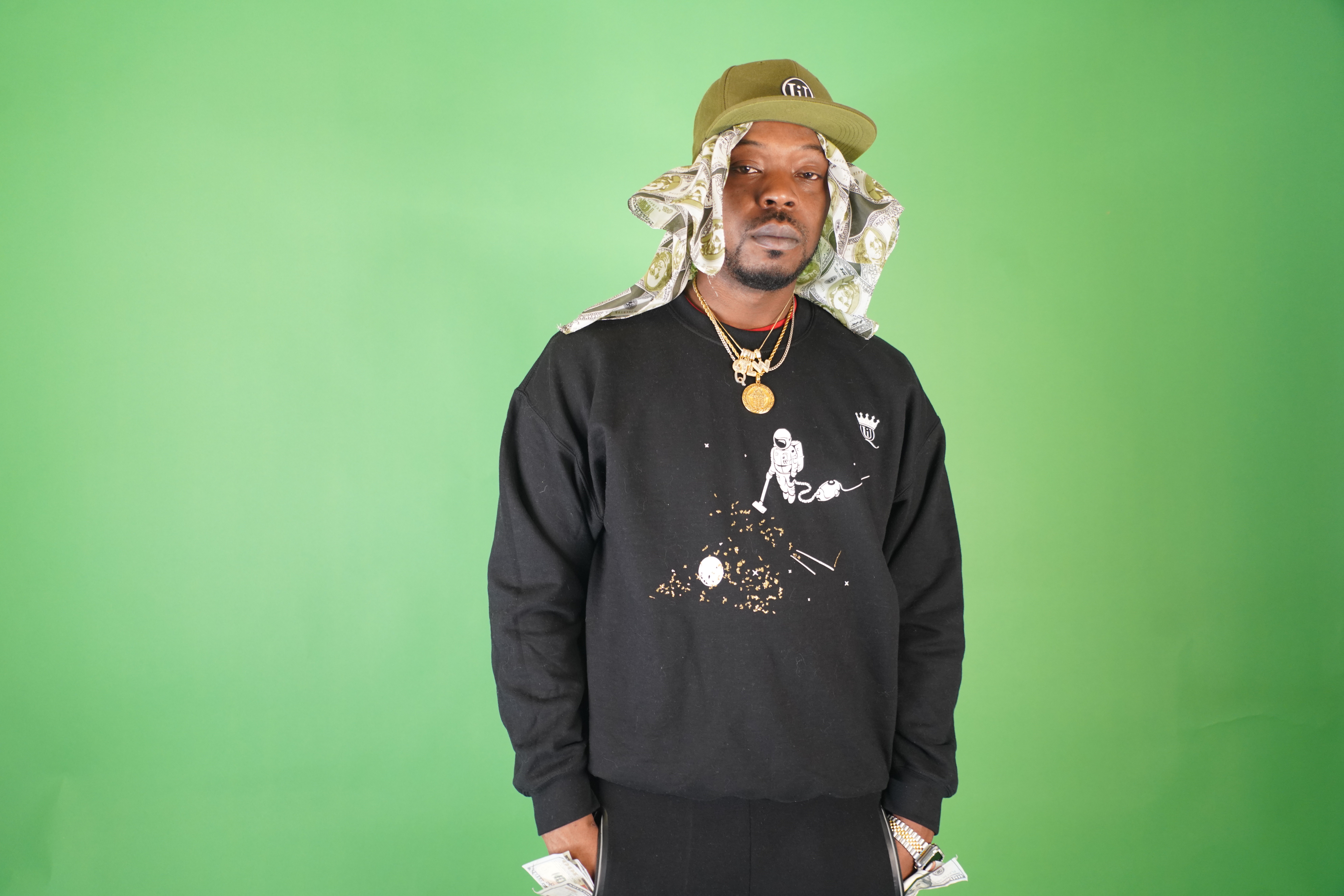Man with long hair wearing a hat, black sweatshirt, and jewelry stands against a green background.