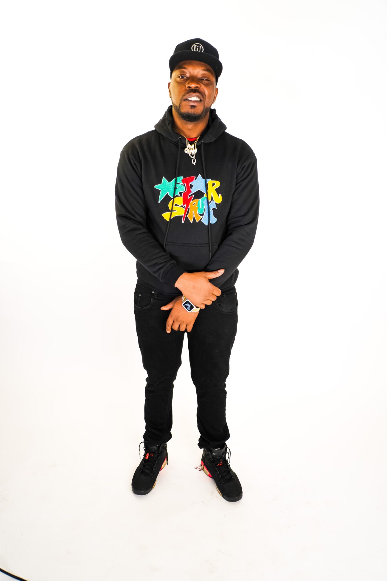 Man standing with arms crossed, wearing a black hoodie with colorful text, black pants, and sneakers, smiling against a white background.