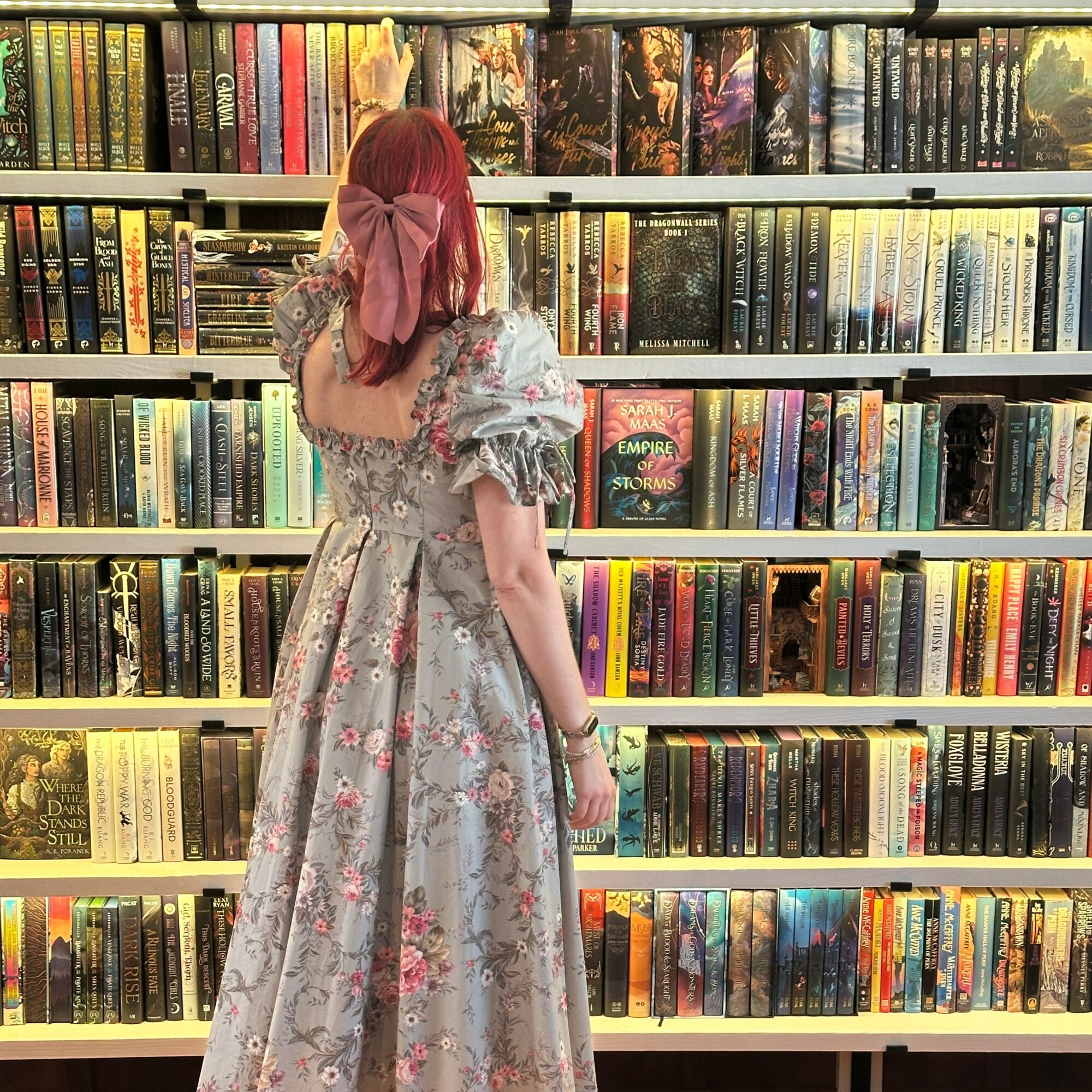 Person with red hair and pink bow looking at books on a shelf in a bookstore, wearing a floral dress.