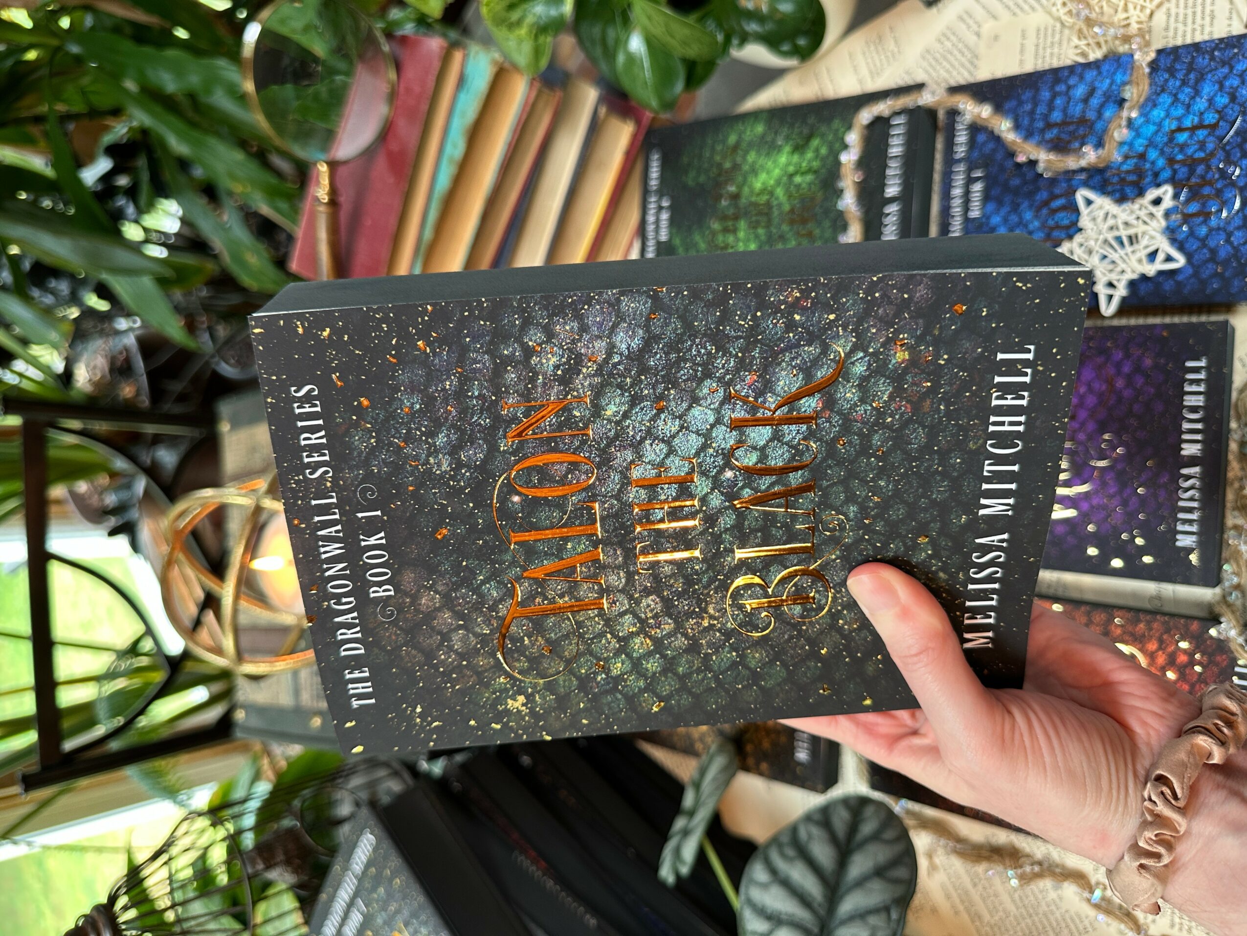 Person holding a book titled 'The Bone Season' by Samantha Shannon, with a background of plants and other books.