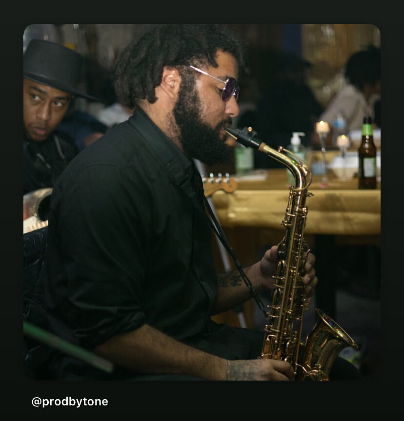 Man playing saxophone in a dimly lit bar with bottles on the counter, wearing glasses and a black shirt.