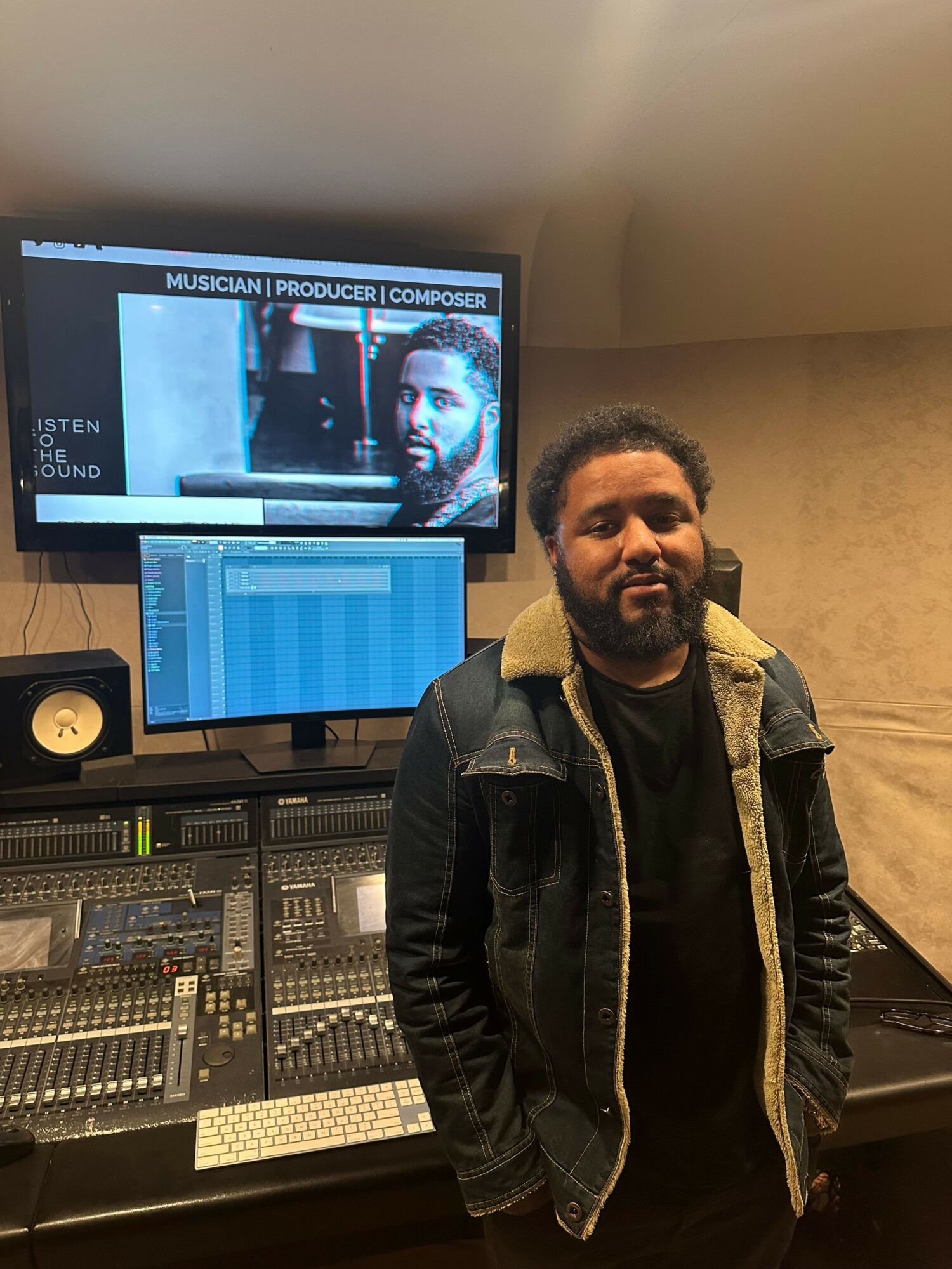 Man standing next to audio mixing console and computer monitor in a recording studio.