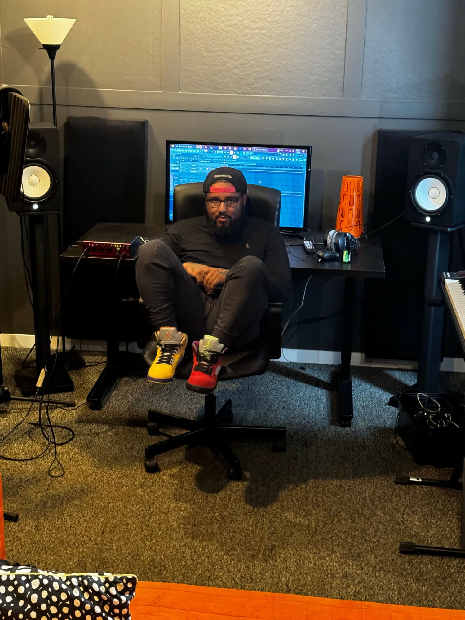 Person sitting at a desk in a recording studio with audio equipment and a computer monitor.