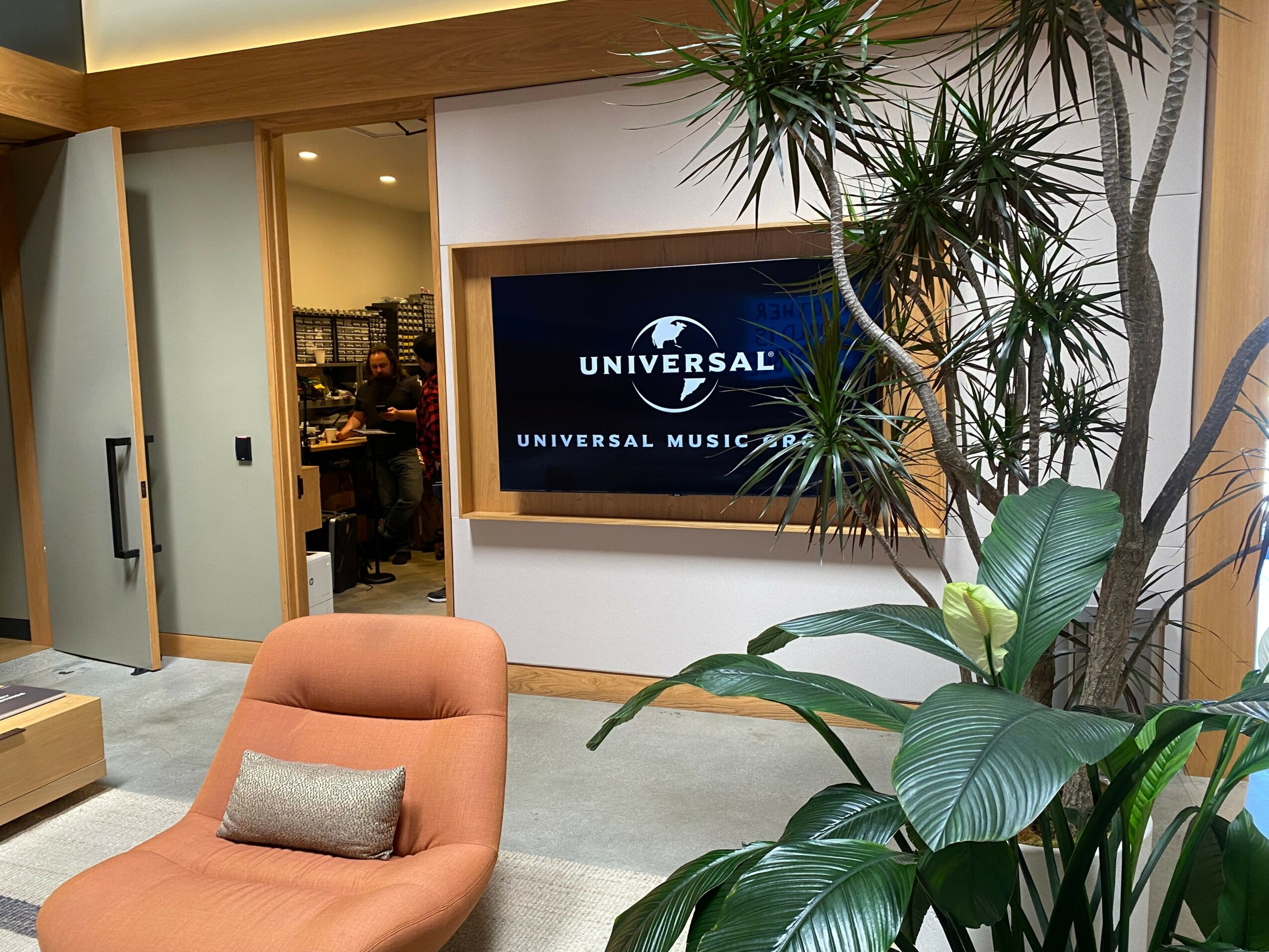 Indoor space with a large plant, a television screen displaying 'Universal Music Group,' and a person visible through a doorway.