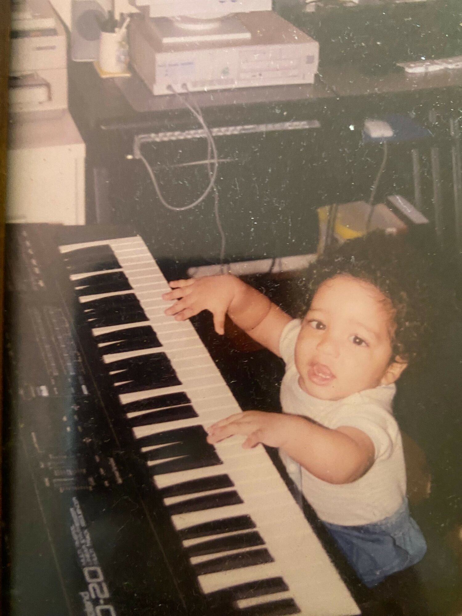 Child leaning over a keyboard, reaching with one hand, in a room with electronic equipment nearby.