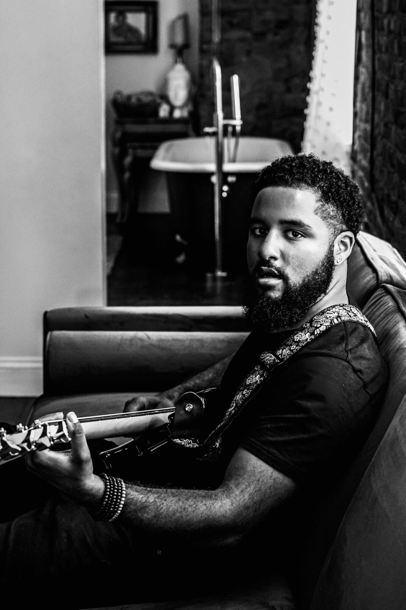 Man with a beard sitting on a couch holding a guitar, in a room with a bathtub in the background.