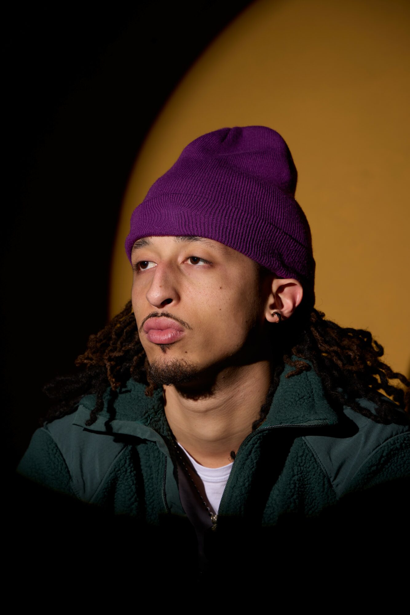 Young man with a purple beanie, curly hair, and a dark jacket, looking to the side against a dark background.