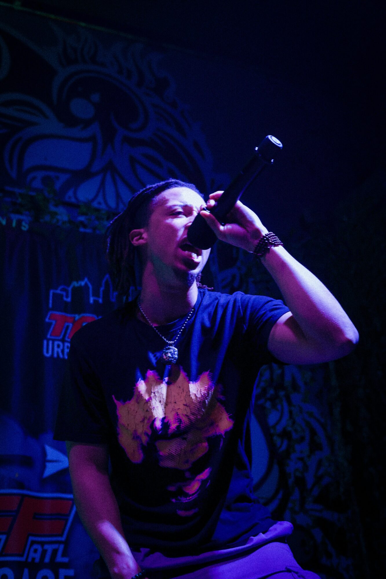 Person singing into microphone on stage with dark background and colorful lighting.