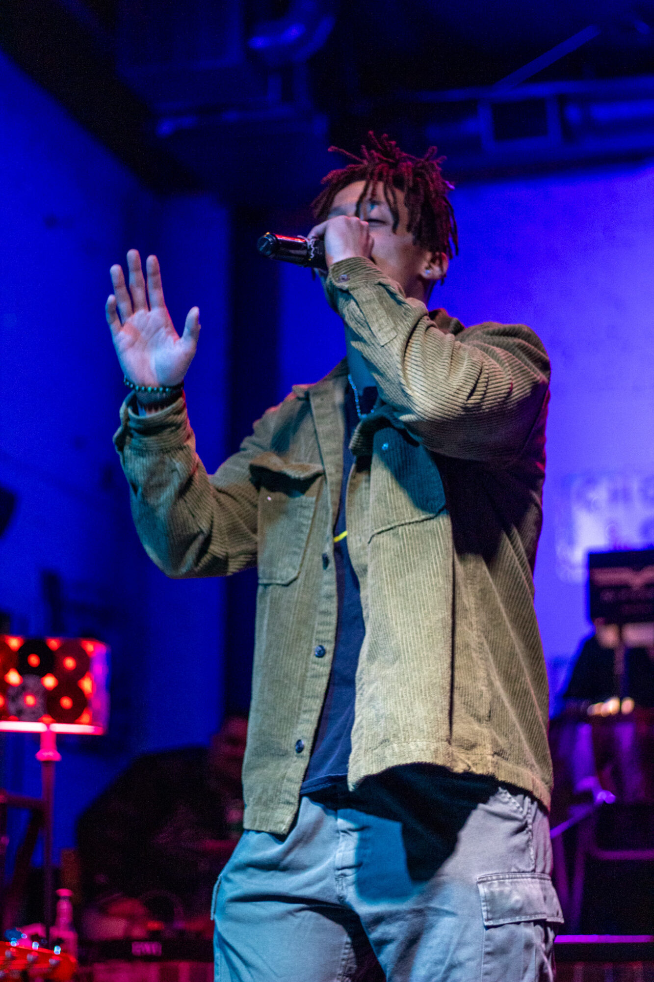 Performer singing into microphone on stage with blue and purple lighting, wearing a brown jacket and gray pants.