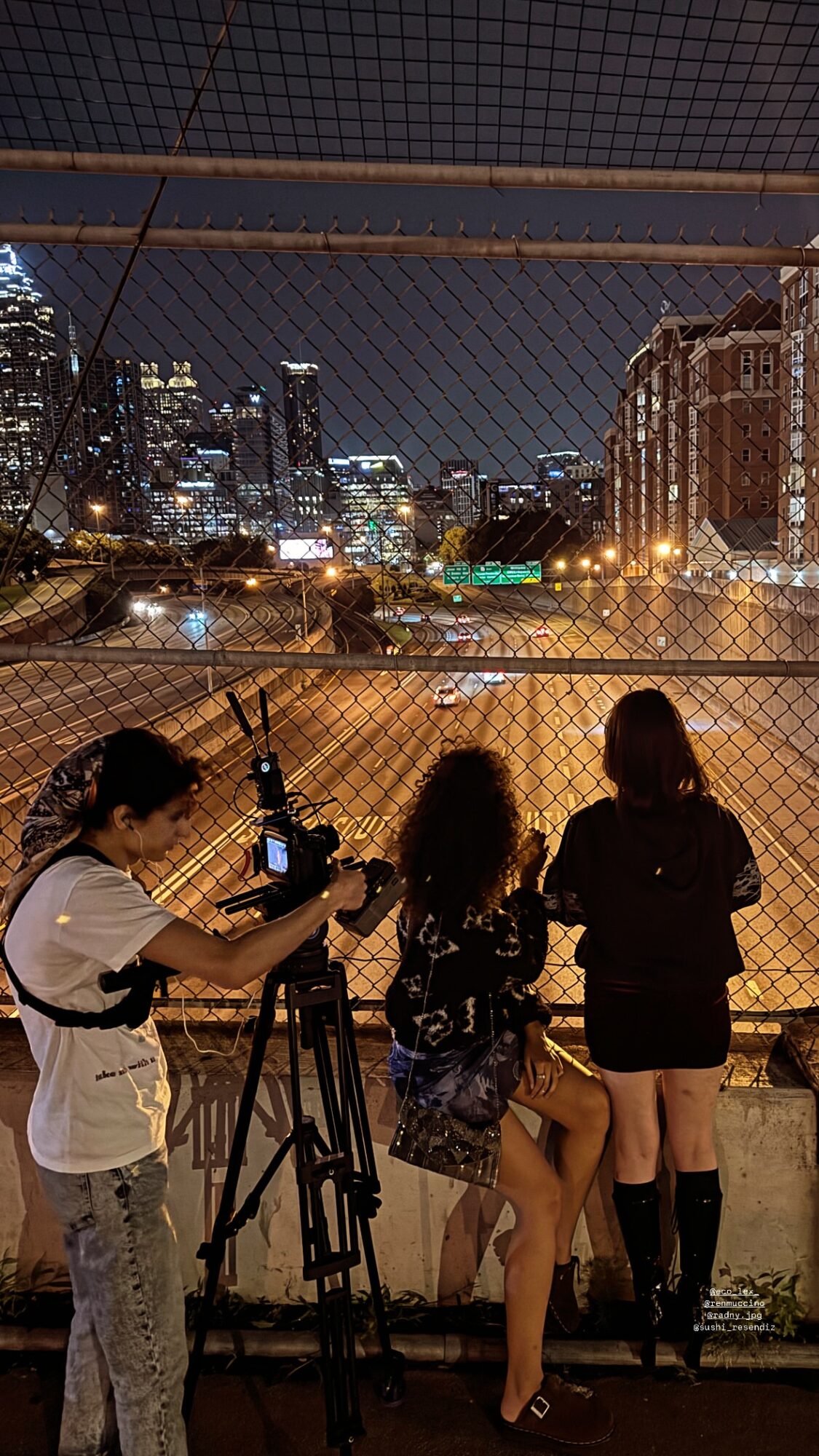 Three women and a person with camera on a tripod watch city skyline at night from a fenced rooftop.