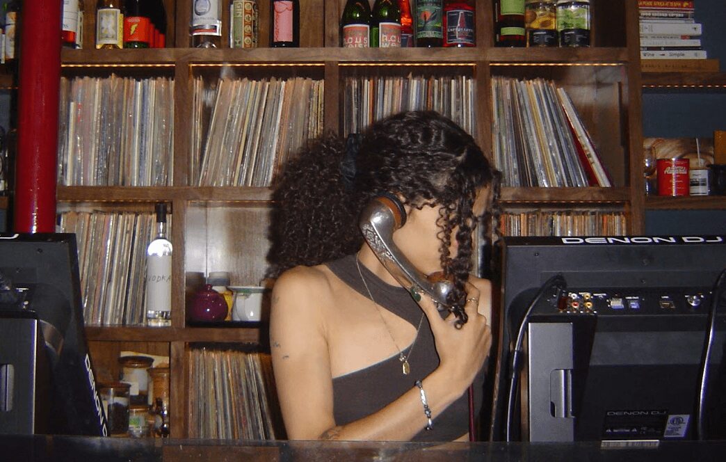 Woman with curly hair talking on a vintage telephone in front of a bookshelf filled with records and bottles.