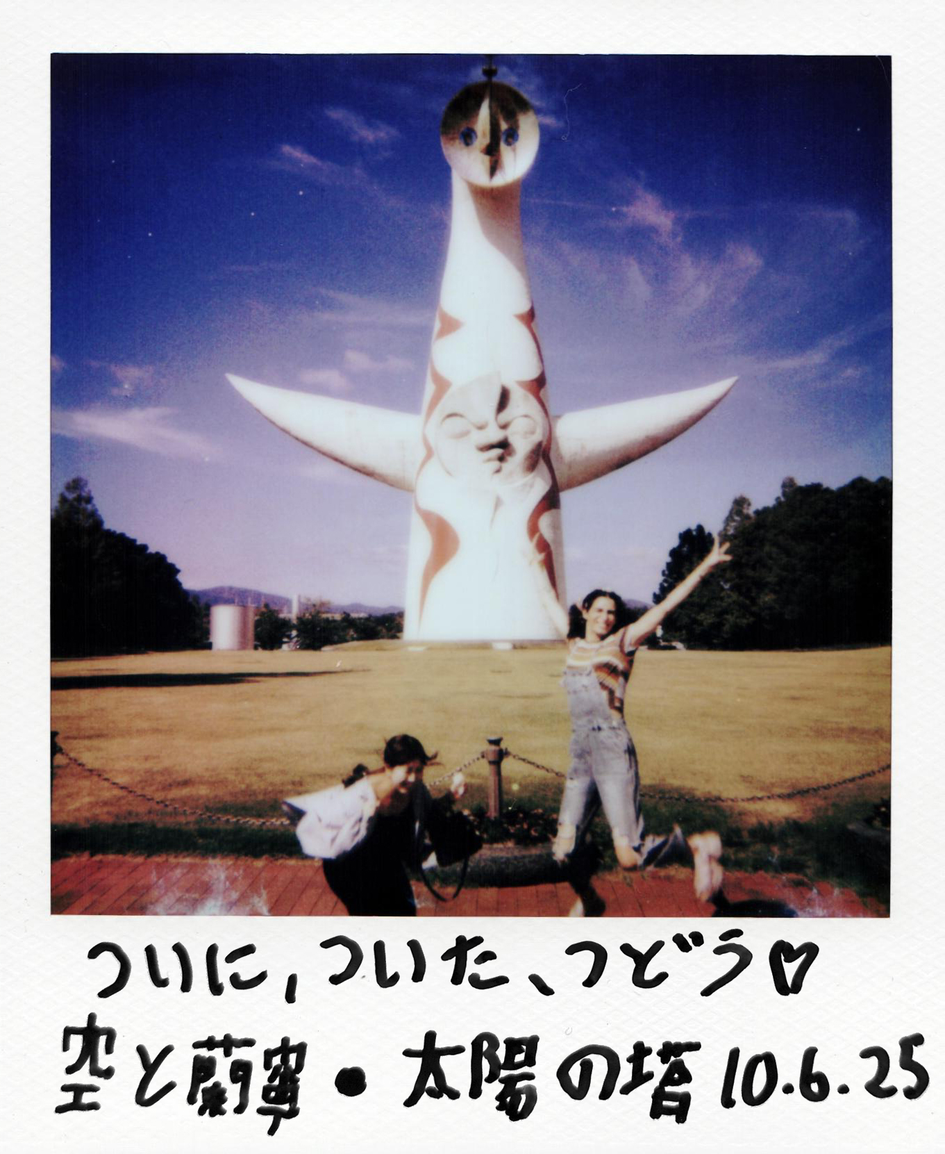 Two people in front of a large giraffe-shaped sculpture with a blue sky background, one person raising an arm, the other kneeling, handwritten text below.