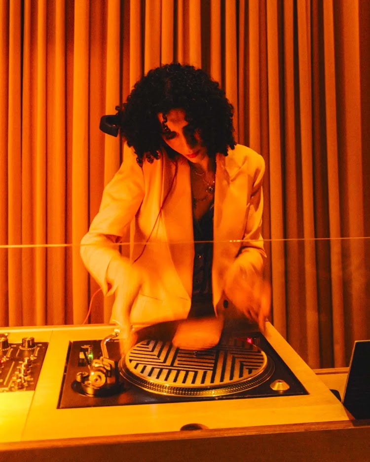 Person with curly hair wearing a light-colored blazer DJing at a turntable, orange curtains in background.
