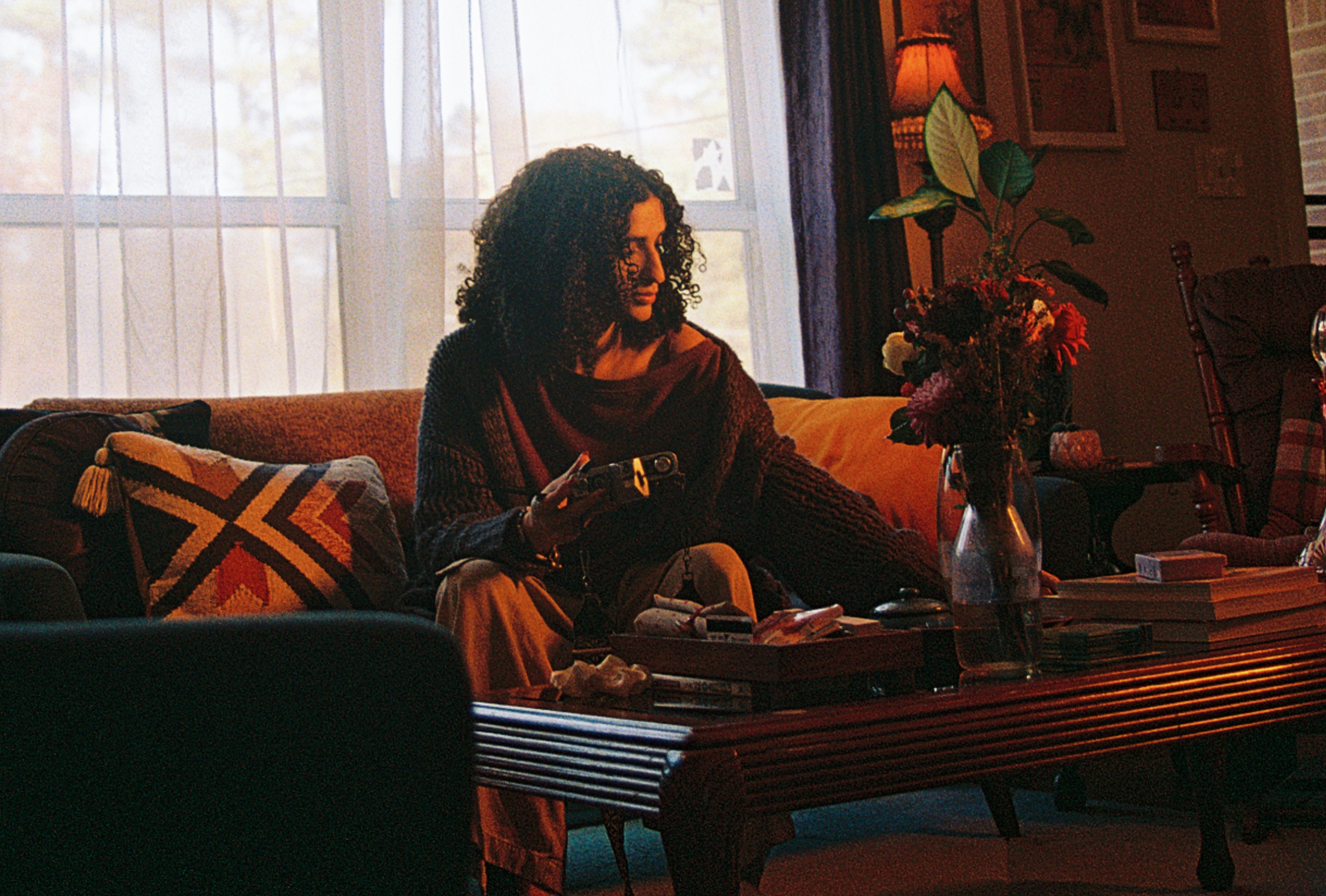 Woman with curly hair sitting on a sofa, holding a phone, in a cozy living room with a coffee table and flowers.