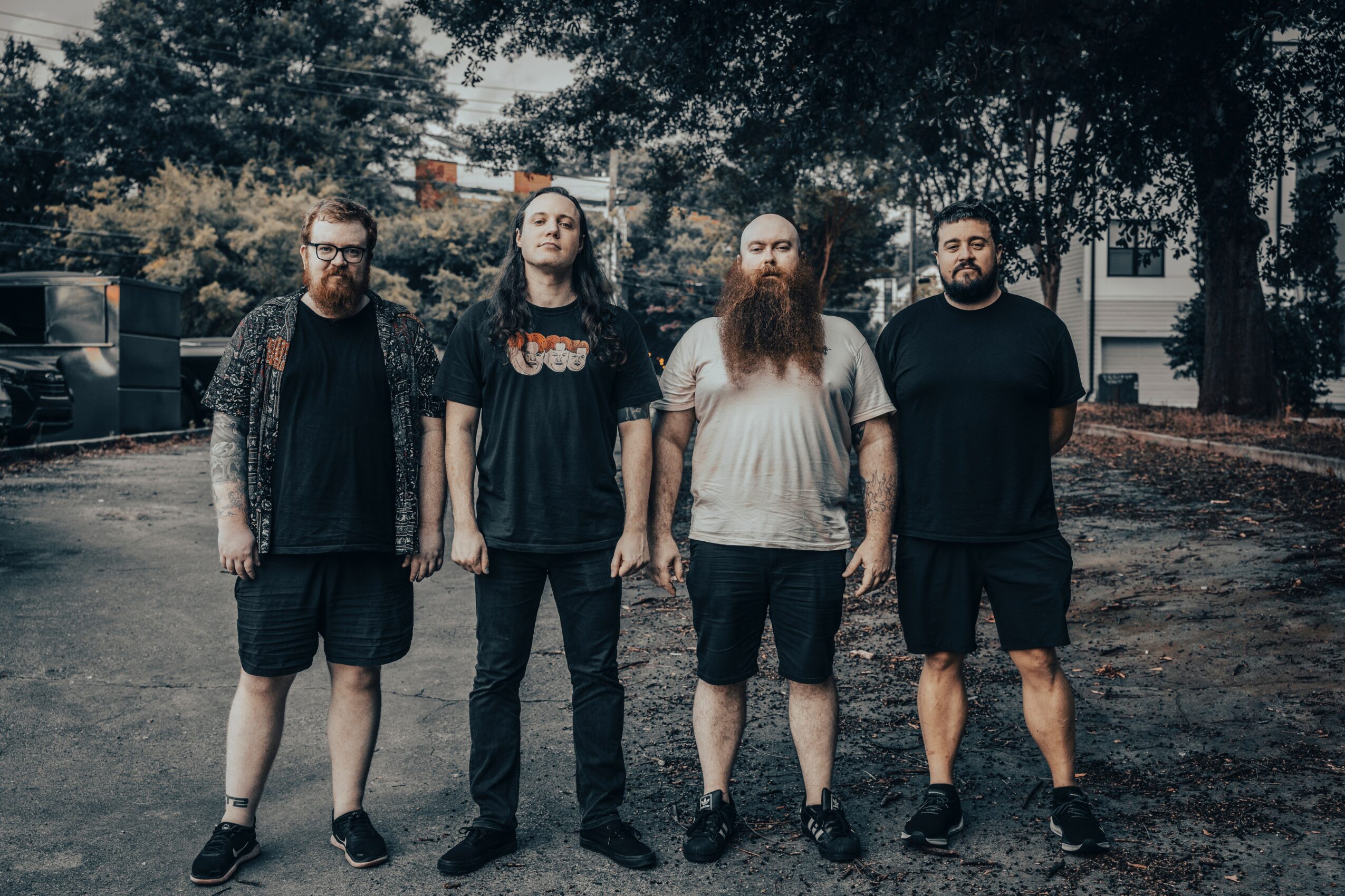 Four men stand outdoors on a paved area with trees and buildings in the background, facing the camera.