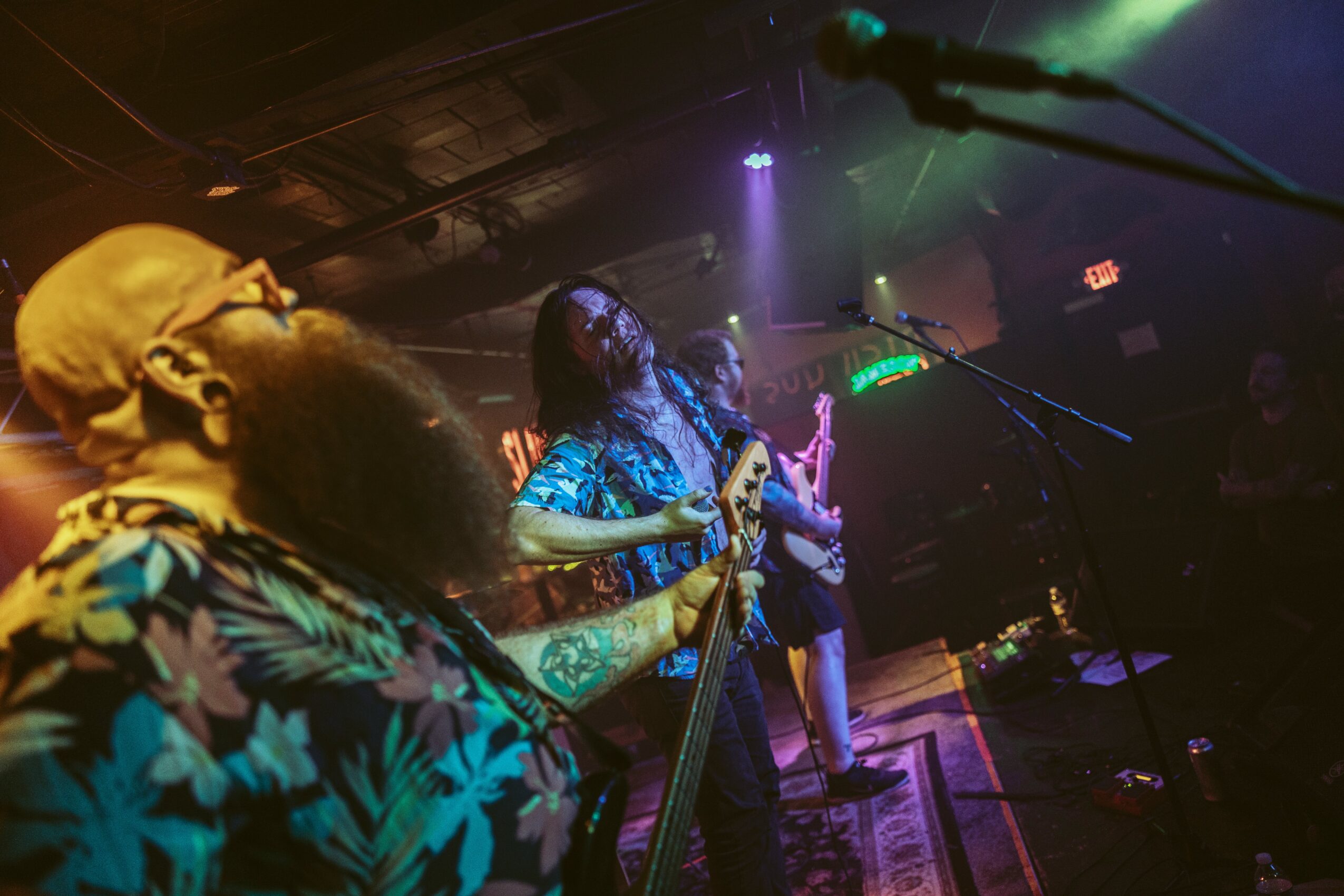 Musicians perform on stage with colorful lighting, one with a beard and glasses, another with long hair, in a dim venue.
