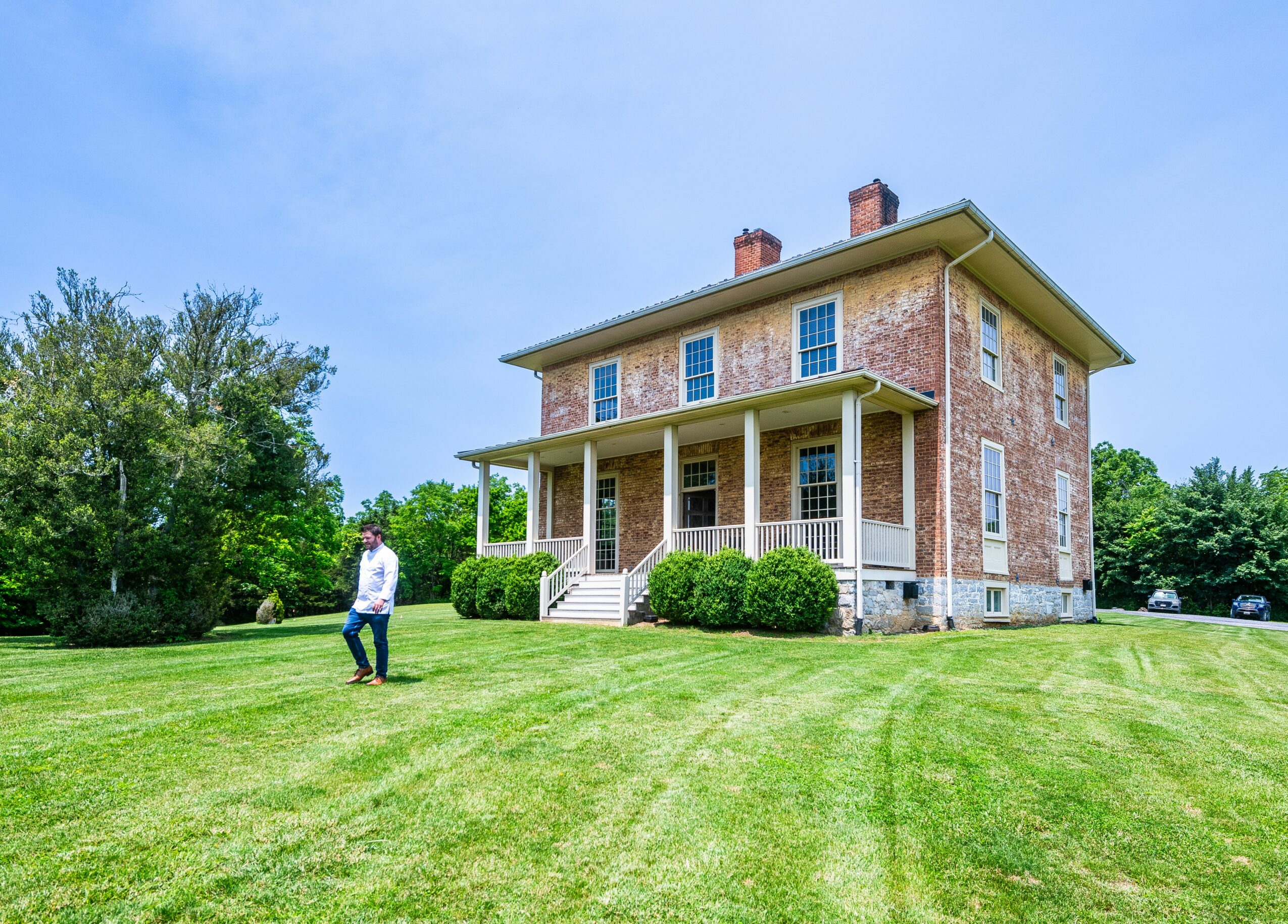 Three-story brick house with a porch, surrounded by green lawn and trees, under a blue sky.