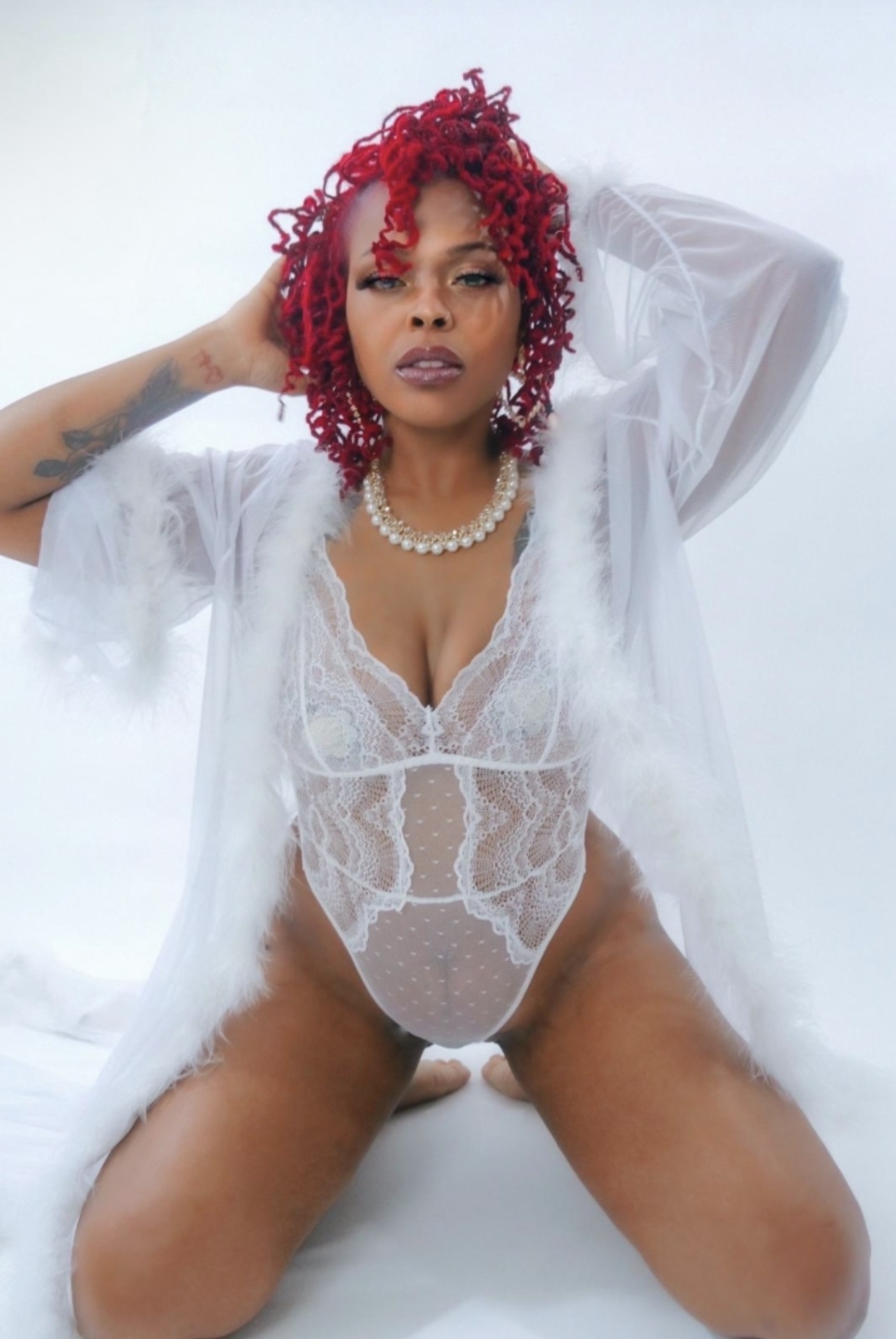 Woman with red curly hair wearing lace bodysuit and pearl necklace, posing with arms raised, against a white background.